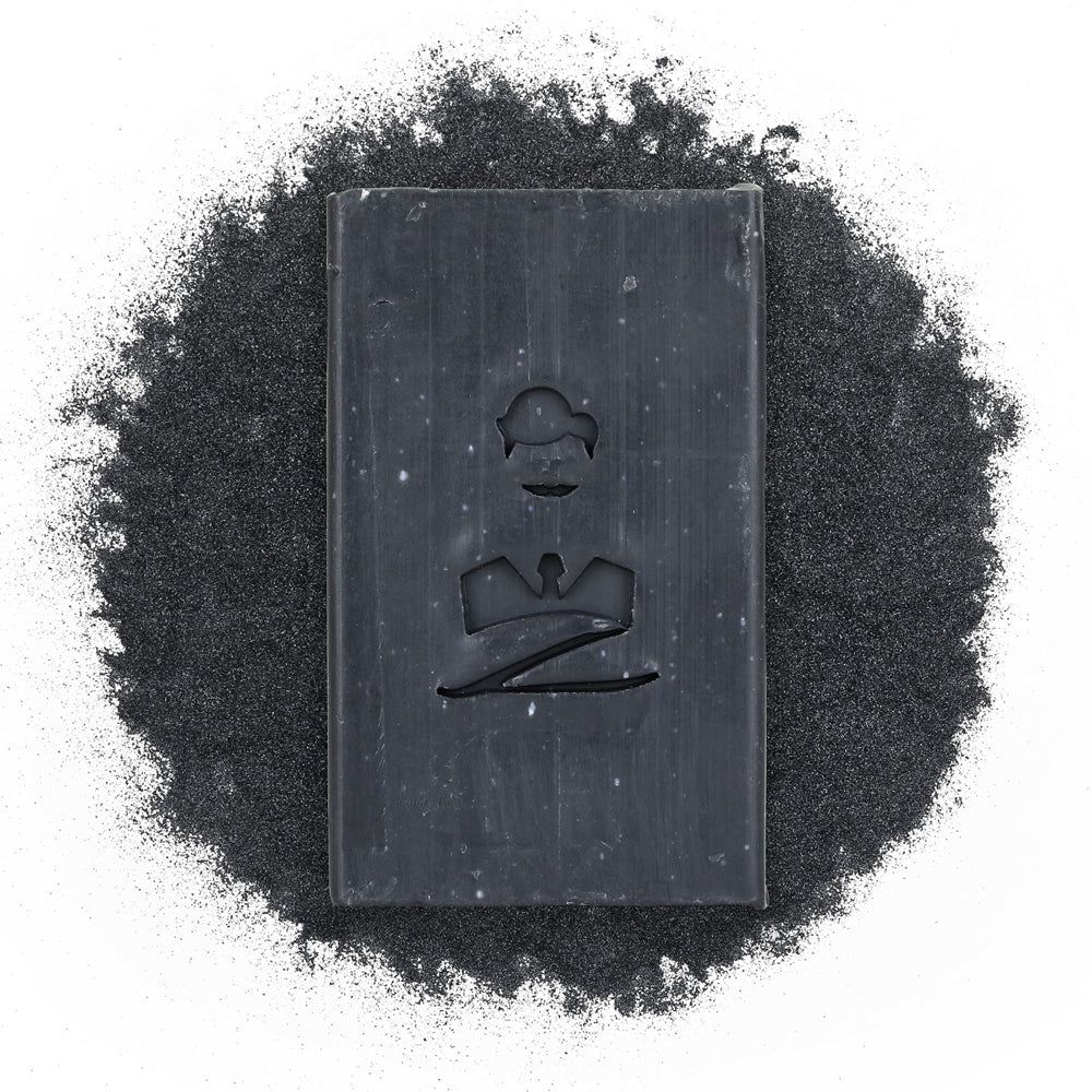 Rechteckige, schwarze Seife mit Logo, umgeben von schwarzem Pulver. Logo: stilisiertes Gesicht.
