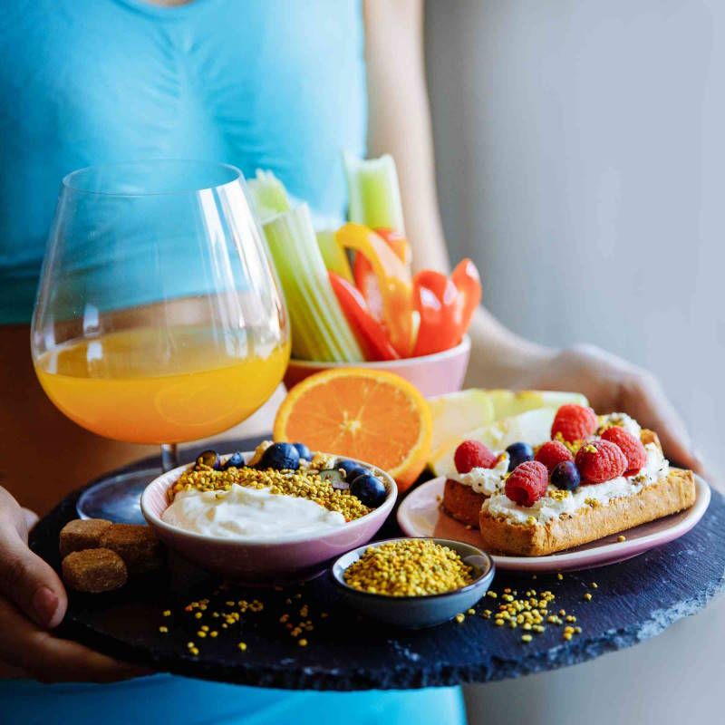 Tablett mit Obst, Gemüse, Joghurt mit Pollen, Toast mit Beeren, Saft im Glas. Person hält Tablett.