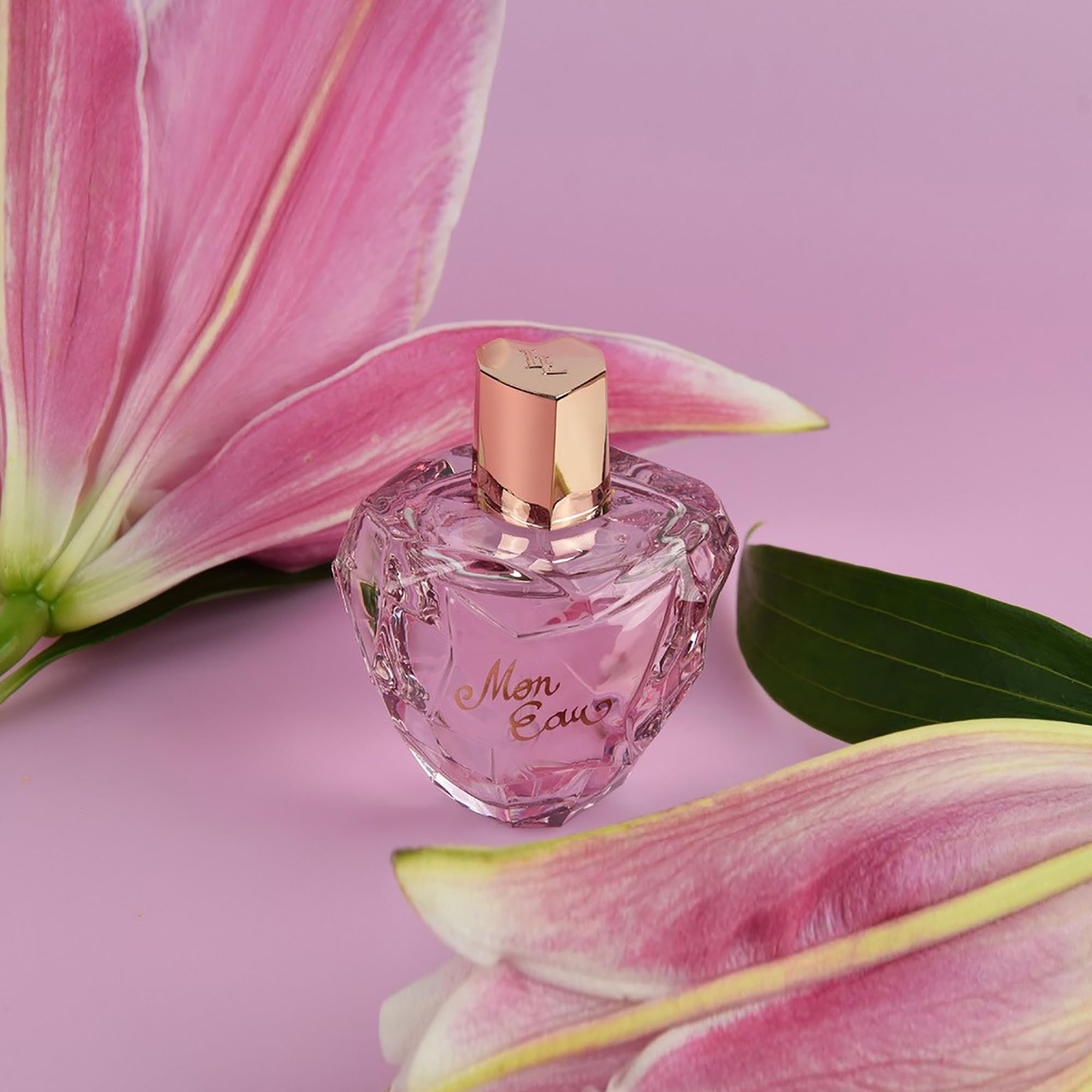 Rosafarbener Flakon mit goldfarbenem Verschluss, umgeben von rosa Lilienblüten. Aufschrift: Mon Eau.
