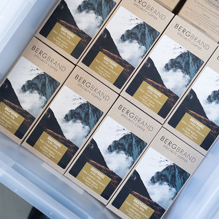 Mehrere Kaffeepackungen mit Bergmotiv. Text: BERGBRAND Specialty Coffee, Ursprung Filter. Verpackungen in einer transparenten Box.