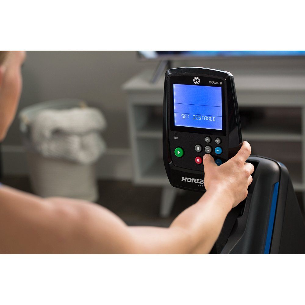 Person hält Bedienfeld. Display zeigt 'SET DISTANCE'. Horizon Fitness Logo. Schwarzes Bedienfeld mit Knöpfen.