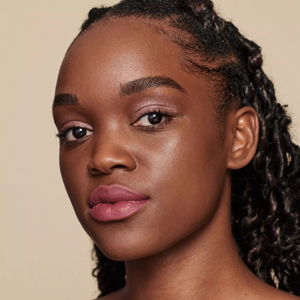 Nahaufnahme einer Frau mit dunkler Haut und Locken. Sie trägt Make-up, einschließlich Lippenstift. Der Hintergrund ist hellbeige.