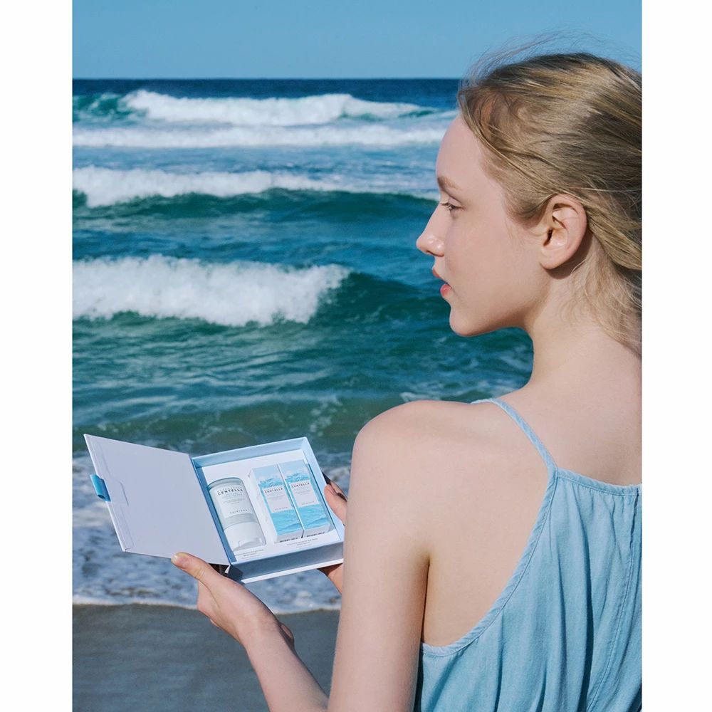 Frau hält eine offene Produktverpackung am Strand. Enthält Sonnenschutzprodukte. Hintergrund: Meer und Himmel.