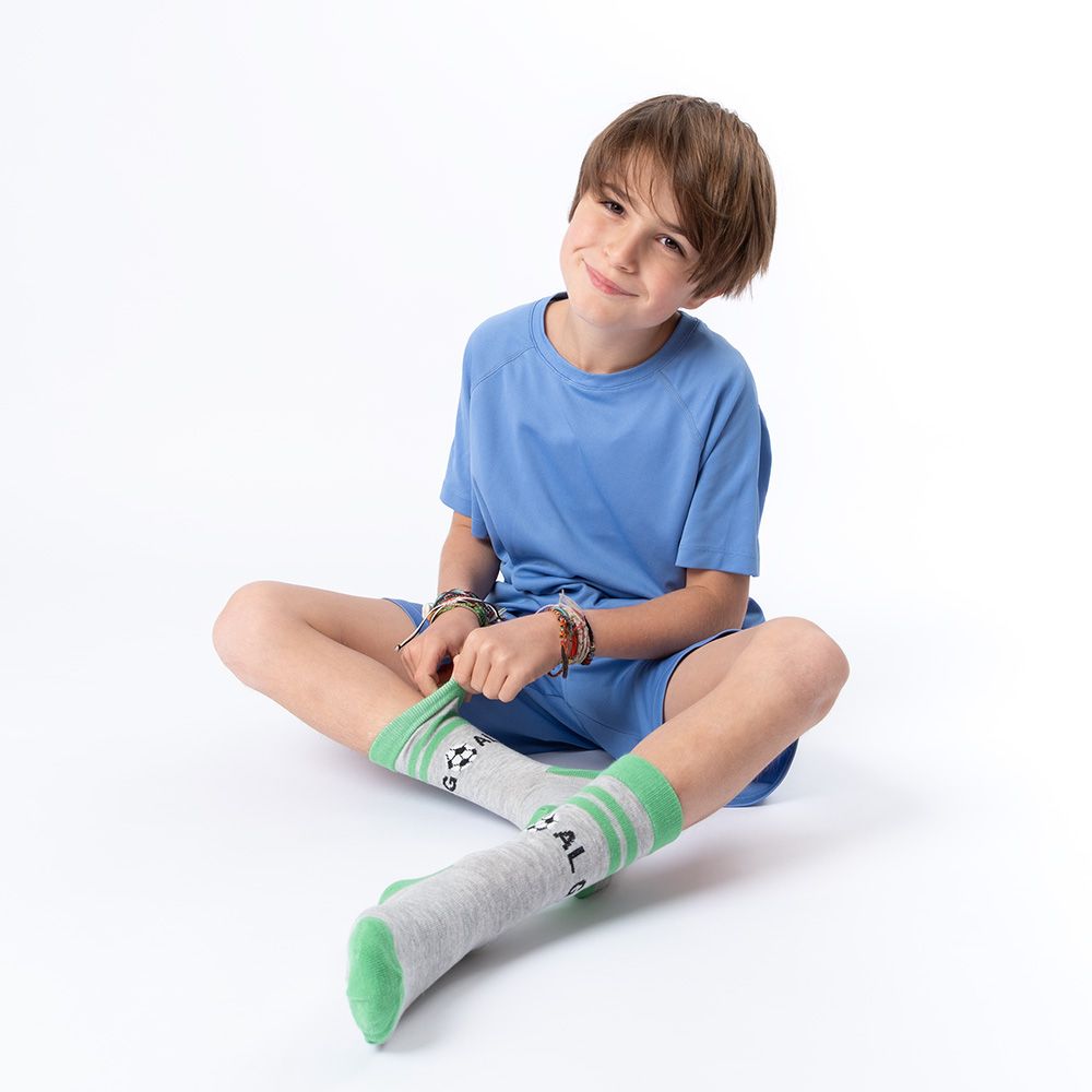 Junge sitzt, zieht Socken an. Graue Socken mit grünem Rand, Fußball-Motiv. Blaue Shorts.
