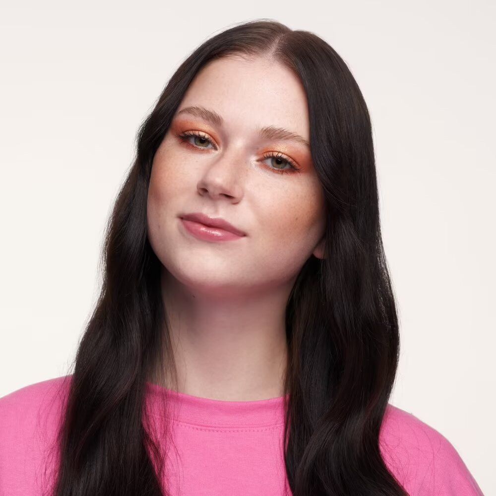 Porträt einer Frau mit Make-up. Sie trägt einen rosa Pullover. Ihr Haar ist dunkel und wellig.