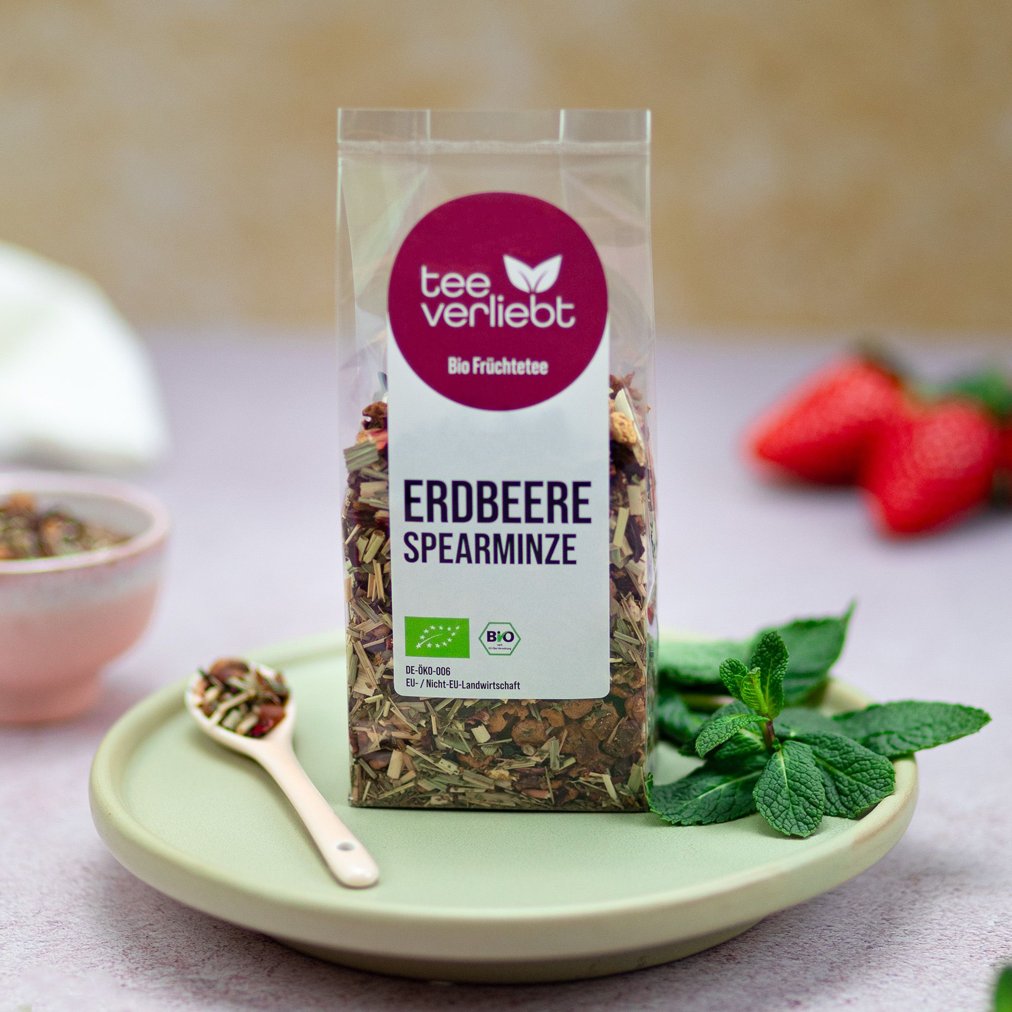 Verpackung mit losem Früchtetee auf einem Teller. Erdbeeren und Minze daneben. Aufkleber mit Logo und Text: teeverliebt, Bio Früchtetee, Erdbeere Spearmintze.