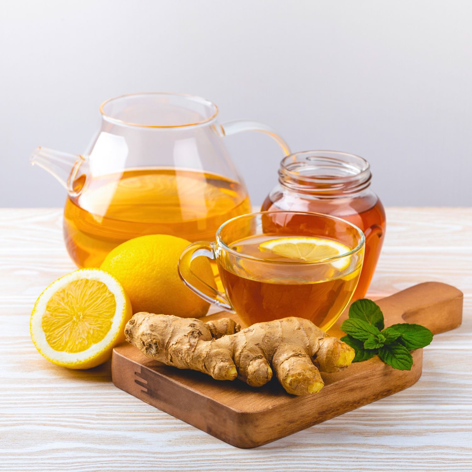 Glas-Teekanne und -Tasse mit Tee, Zitrone, Honig, Ingwer und Minze auf Holzbrett. Auf hellem Untergrund.