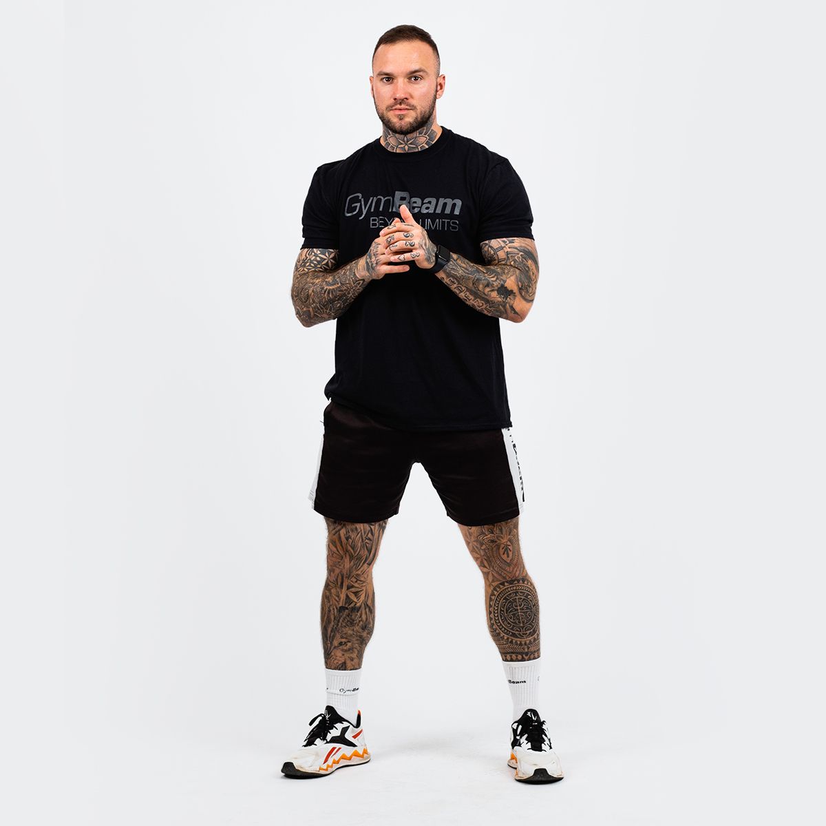Mann in schwarzem "GymBeam BEYOND LIMITS" T-Shirt und kurzen Hosen, steht vor weißem Hintergrund.