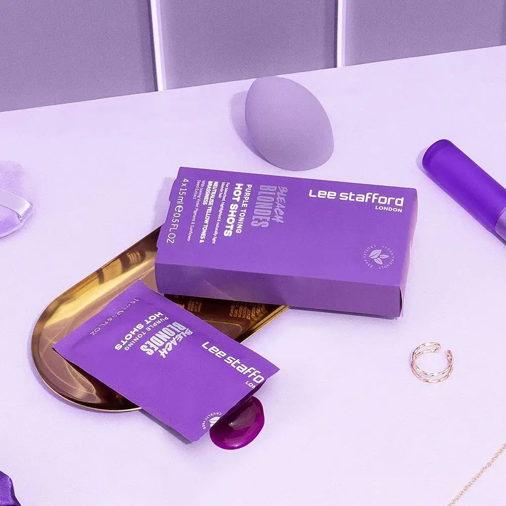 Lila Produktverpackung und Beutel mit Produkt. Goldene Schale, Kosmetik-Applikator und Ring. Lee Stafford Bleach Blondes.