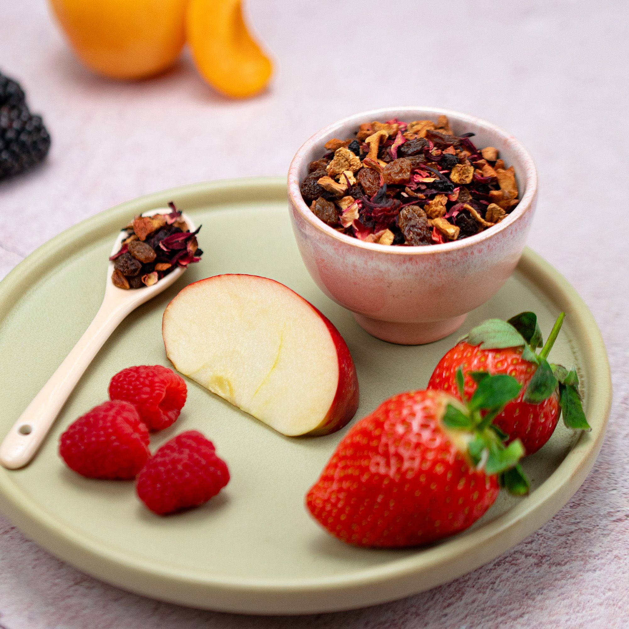 Schale und Löffel mit Teemischung, daneben Erdbeeren, Himbeeren und Apfelstücke auf Untersetzer. 