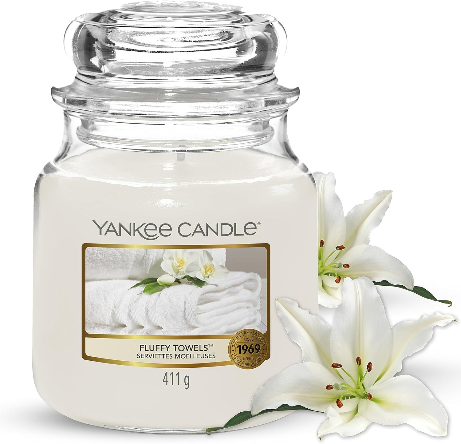 Glasbehälter mit weißem Wachs und Deckel. Etikett mit Text und Bild von Handtüchern und Blumen. "Fluffy Towels" und "Yankee Candle". Lilien.
