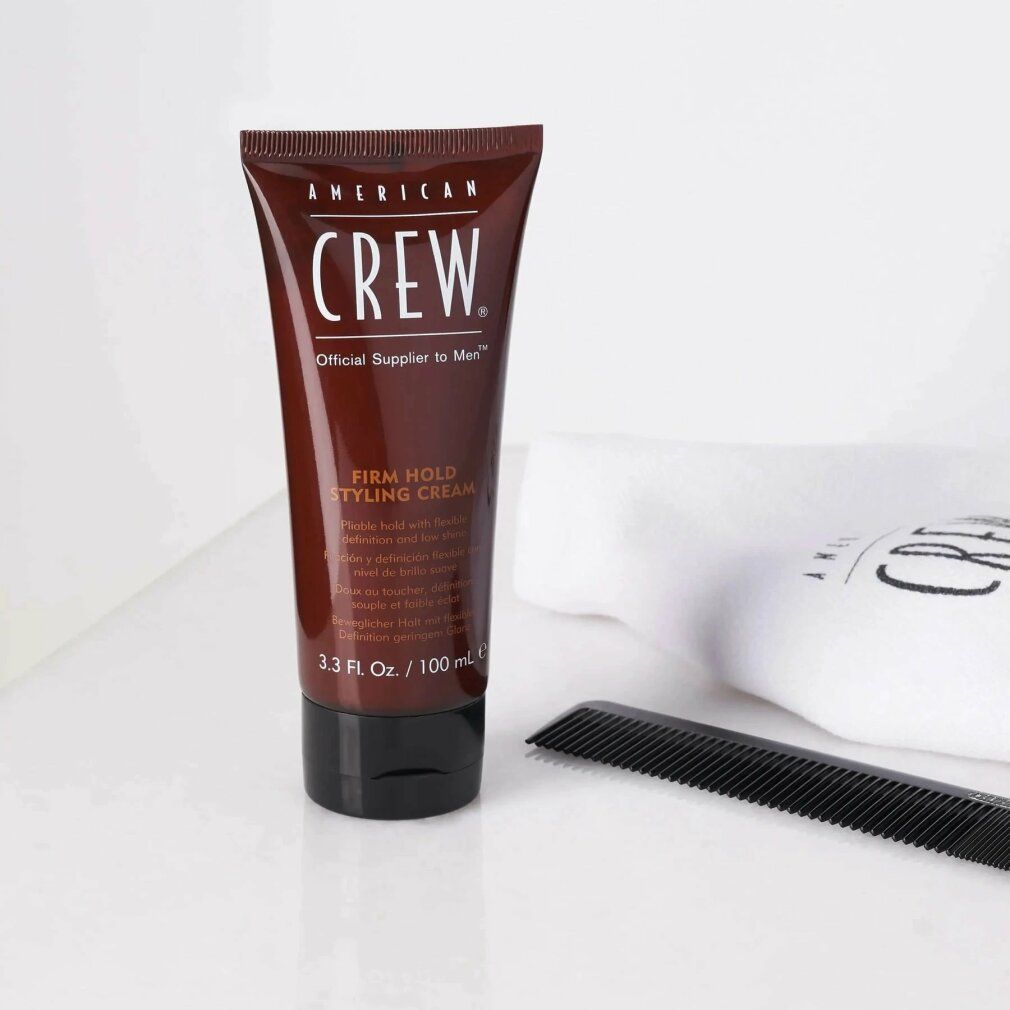 Braune Tube mit schwarzem Deckel. Aufschrift: American Crew, Firm Hold Styling Cream. Daneben Kamm und Handtuch.