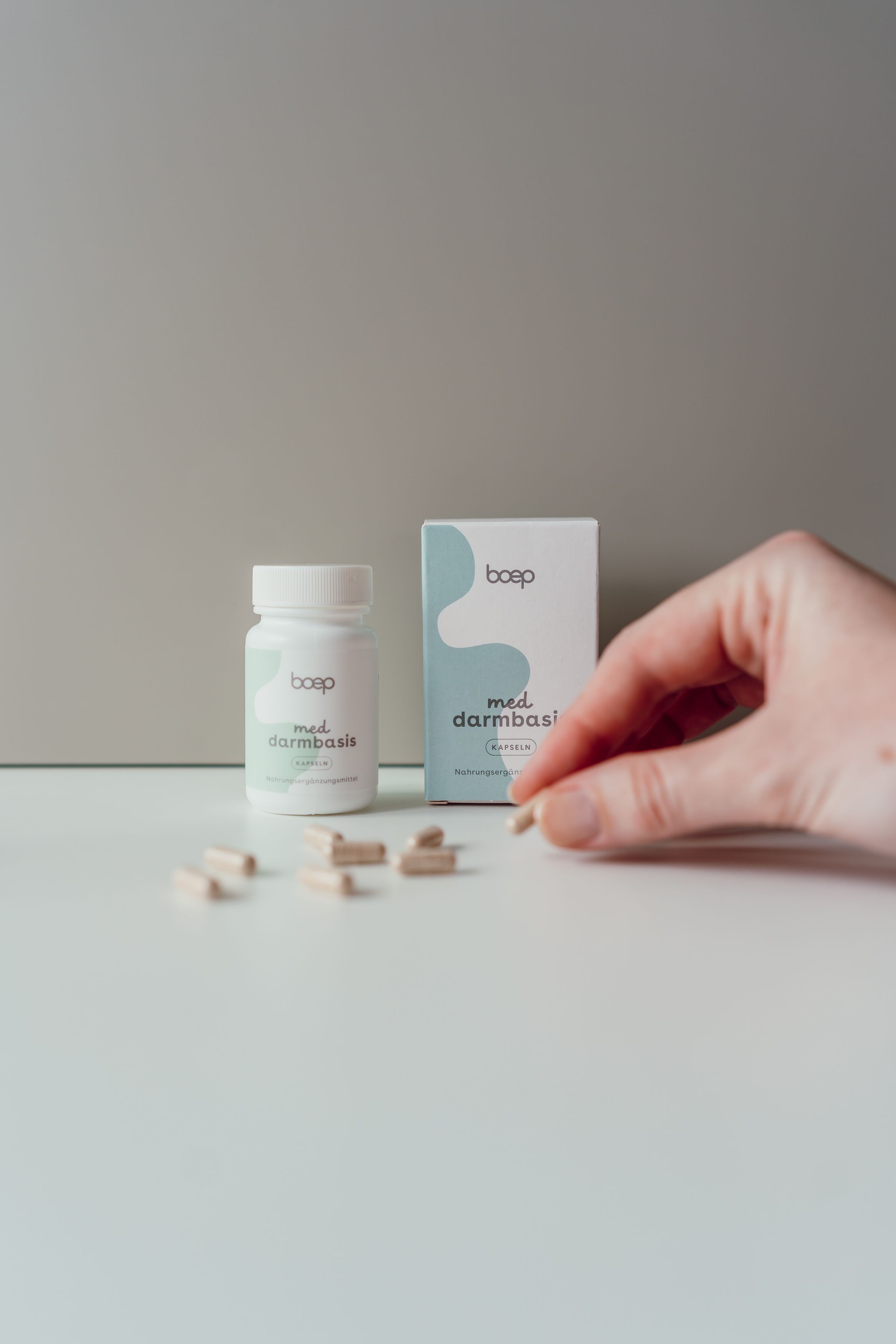 Weiße Flasche und Schachtel mit „boep med darmbasis“ Aufschrift. Kapseln liegen verstreut, Hand greift nach Kapsel.