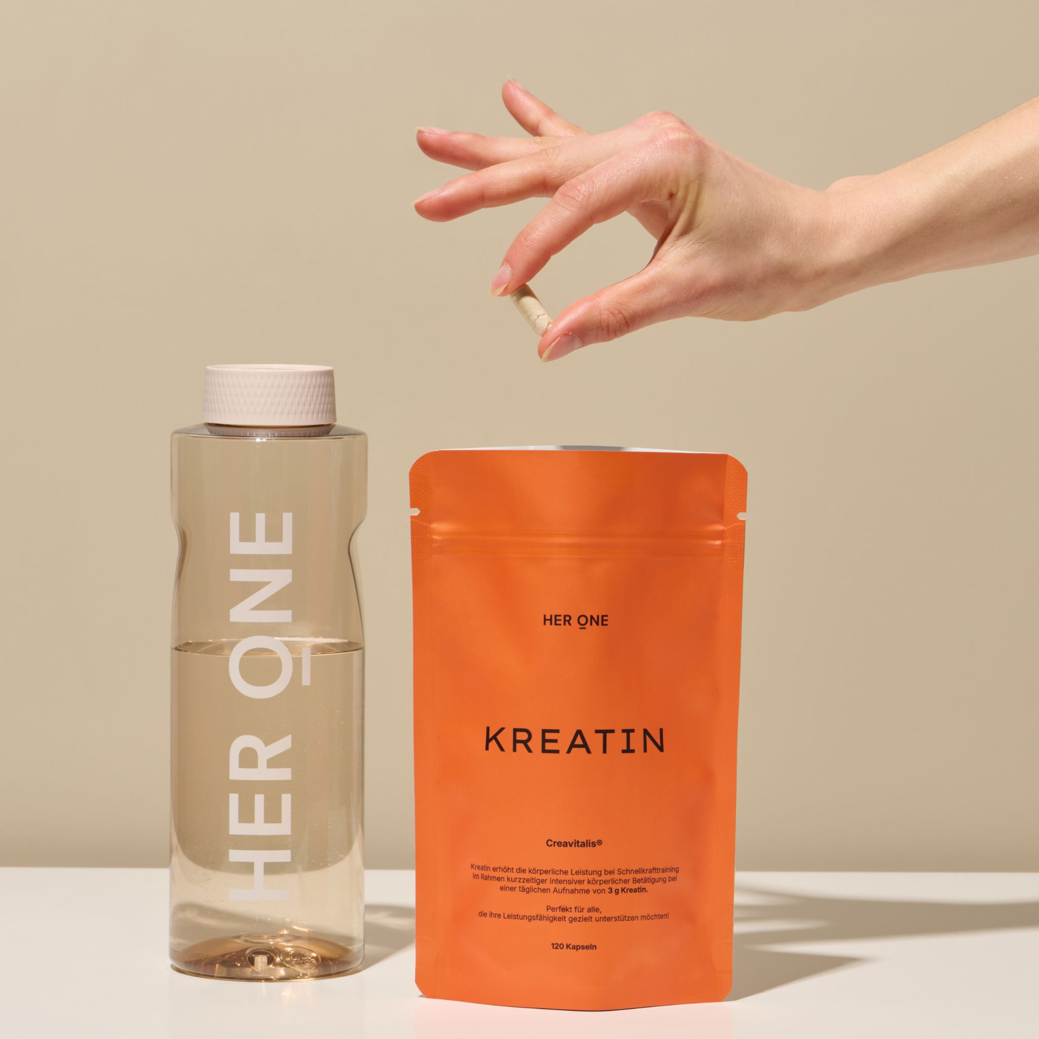 Orangefarbene Verpackung "HER ONE KREATIN" neben einer Wasserflasche und einer Kapsel in einer Hand.
