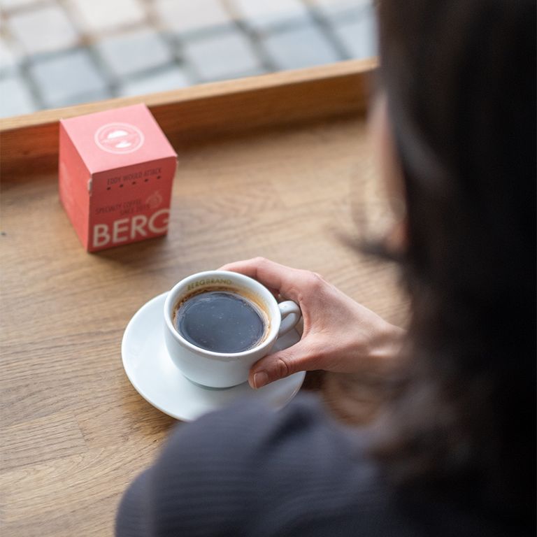 Person hält eine Tasse Kaffee. Auf dem Tisch steht eine rote Verpackung. Aufdruck: BERGBRAND, Kapselkaffee.