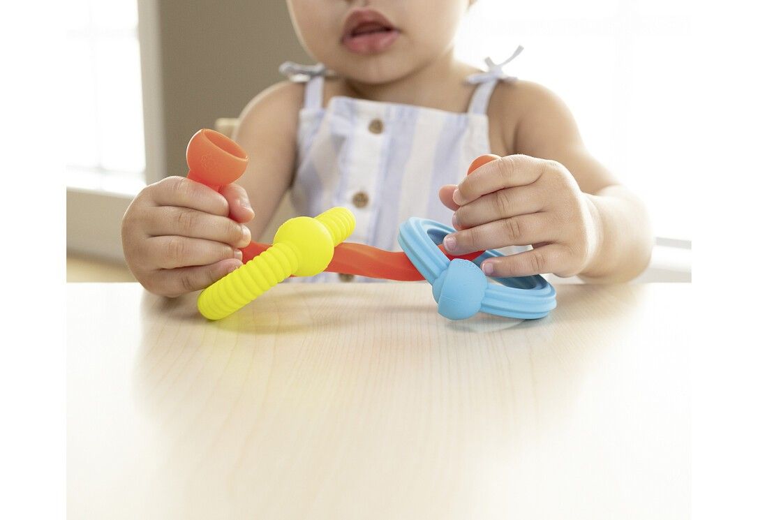 Kind hält farbige Silikonringe. Gelb, rot und blau. Ringe mit Noppen und abgerundeten Enden. Auf hellem Tisch.