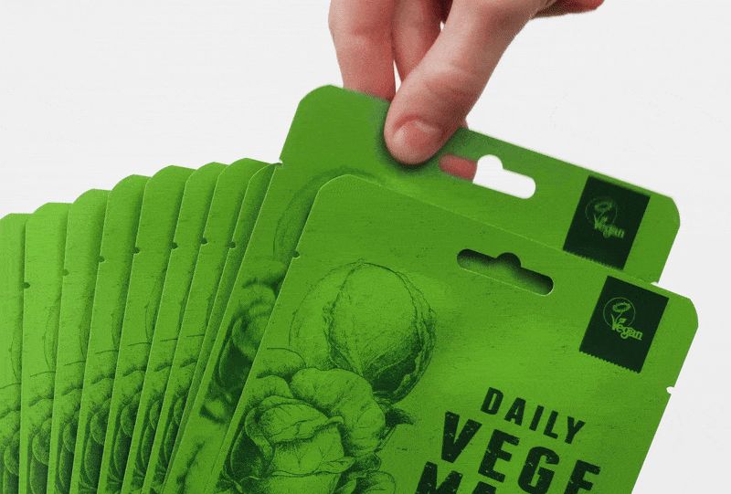 Mehrere grüne Gesichtsmasken mit Aufdruck "DAILY VEGE MASK". Eine Hand greift nach einer Maske. Vegan-Logo.