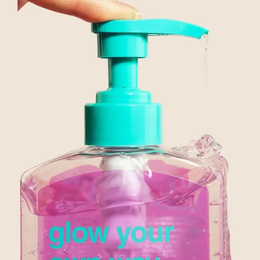 Nahaufnahme einer transparenten Flasche mit rosa Flüssigkeit. Blauer Pumpkopf. Flüssigkeit tropft. Aufschrift: glow your own way.