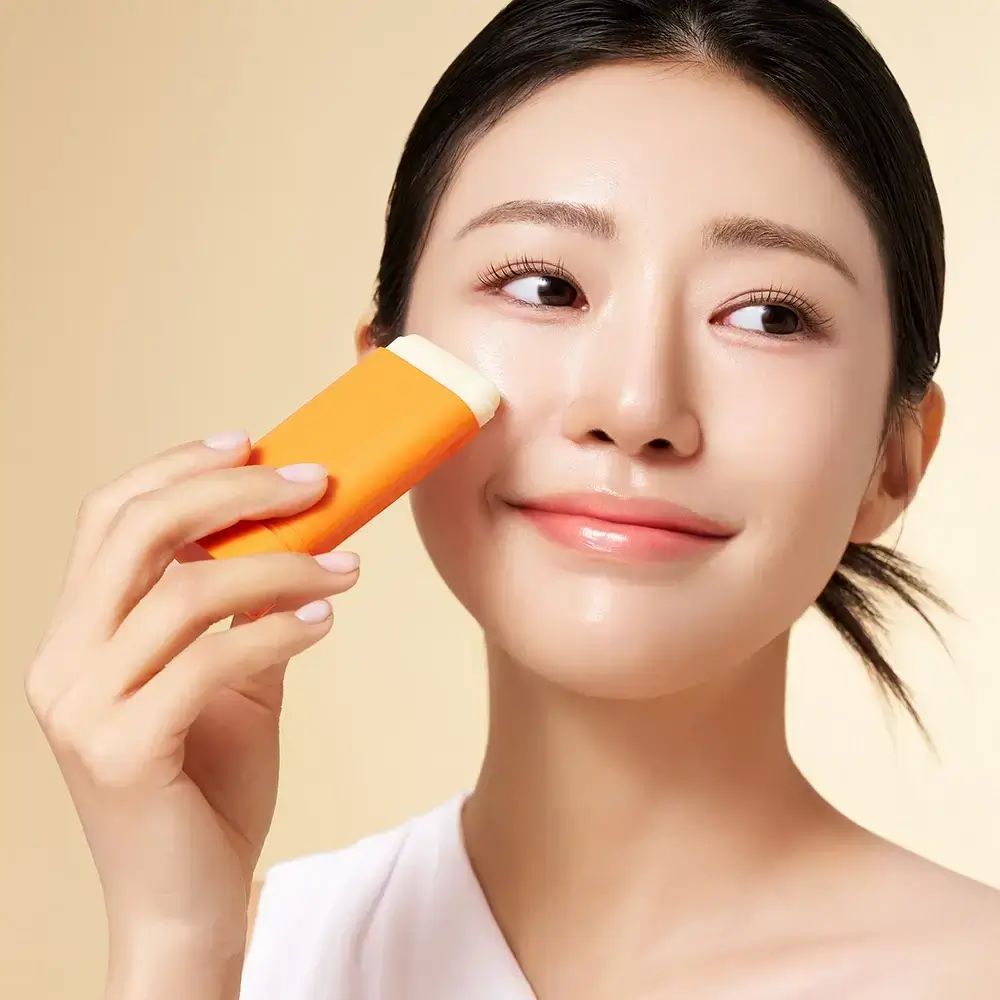Frau trägt orangefarbenen Sonnenschutz-Stick auf die Wange auf. Der Stick hat einen weißen Kopf. Hintergrund: hell.