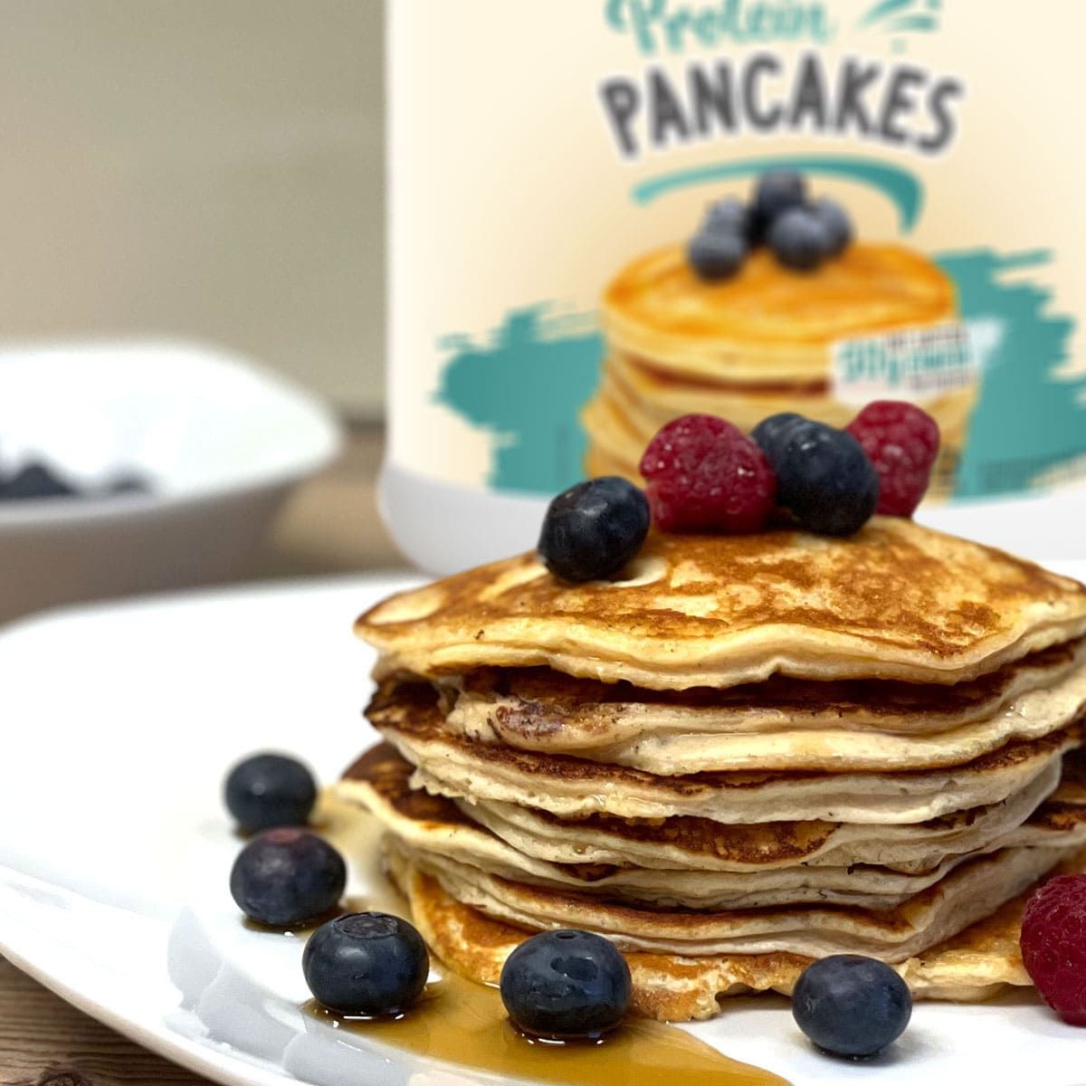 Teller mit gestapelten Pancakes, belegt mit Blaubeeren und Himbeeren. Im Hintergrund: Behälter mit TNT Protein Pancakes. Blaubeeren auf dem Teller.