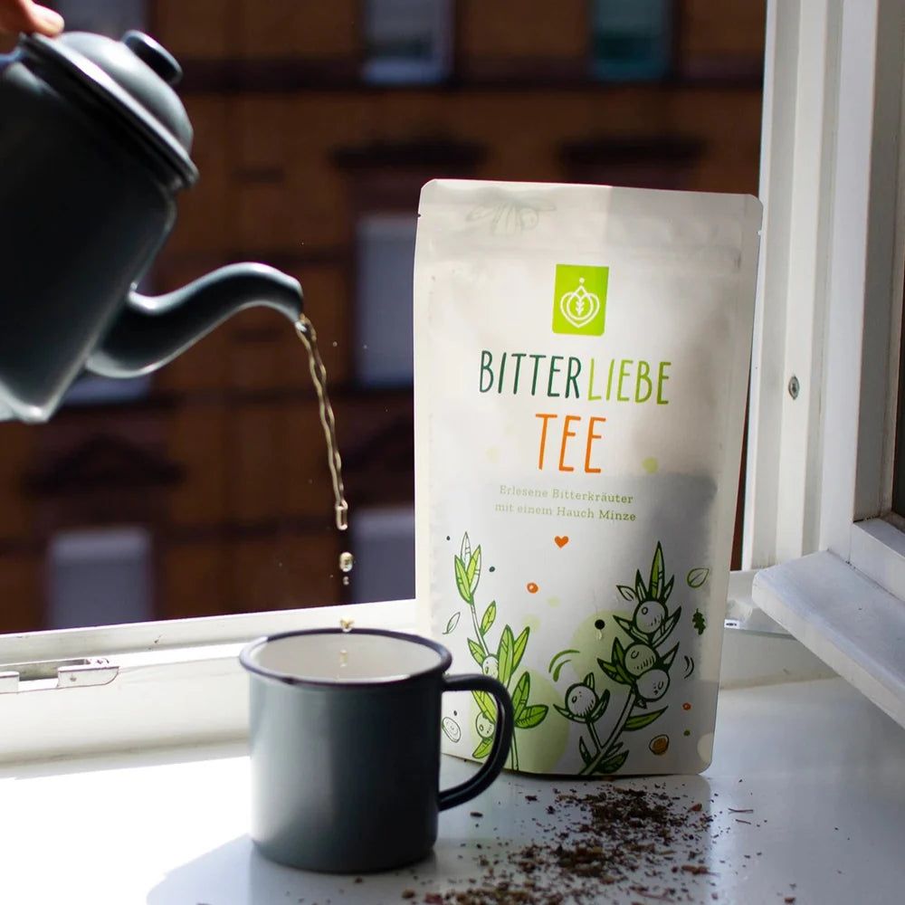Bitterliebe Tee-Beutel neben einer Tasse Tee. Tee wird aus einer Kanne gegossen. Fenster im Hintergrund.