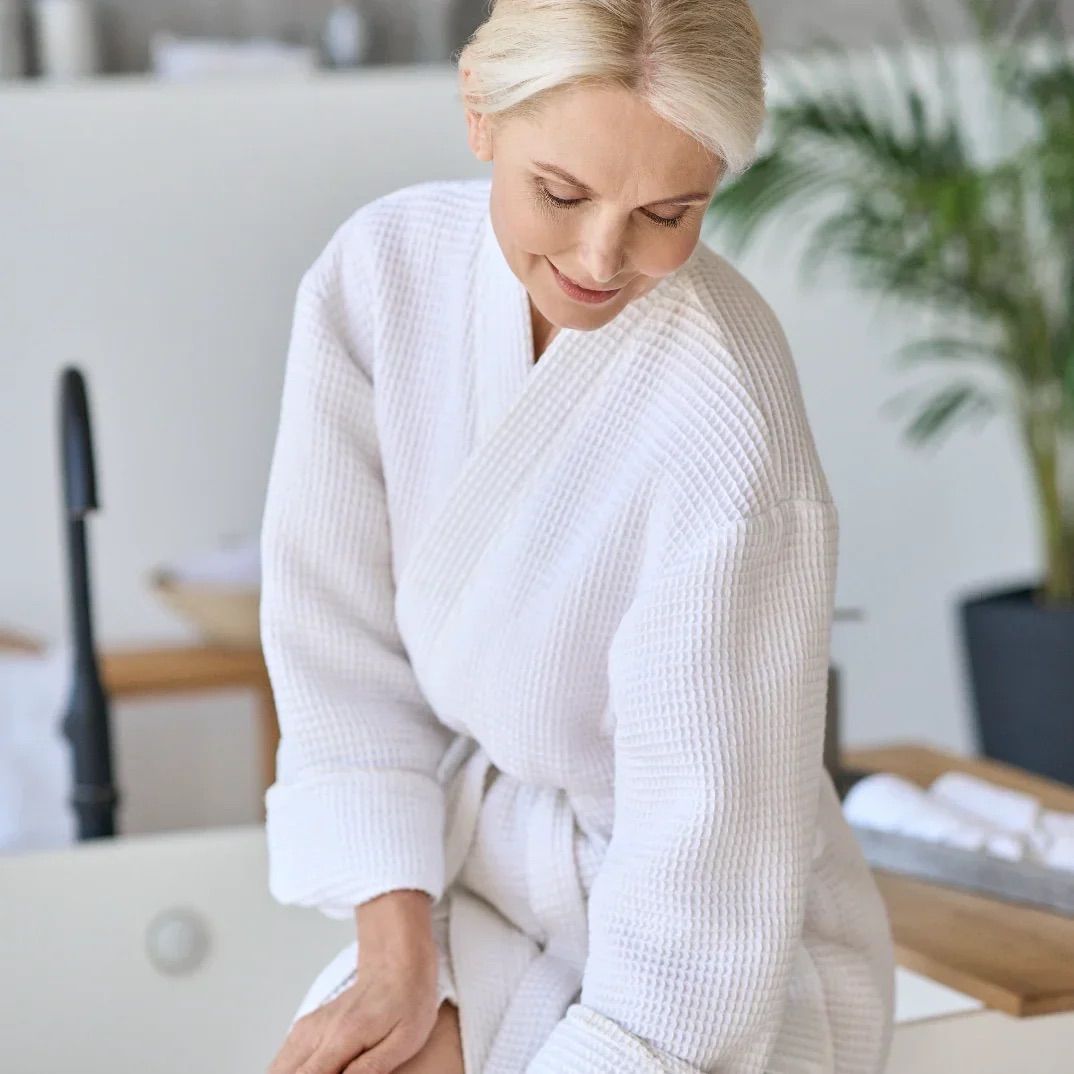 Frau in weißem Bademantel sitzt. Blick nach unten. Hintergrund: Bad, Pflanzen.