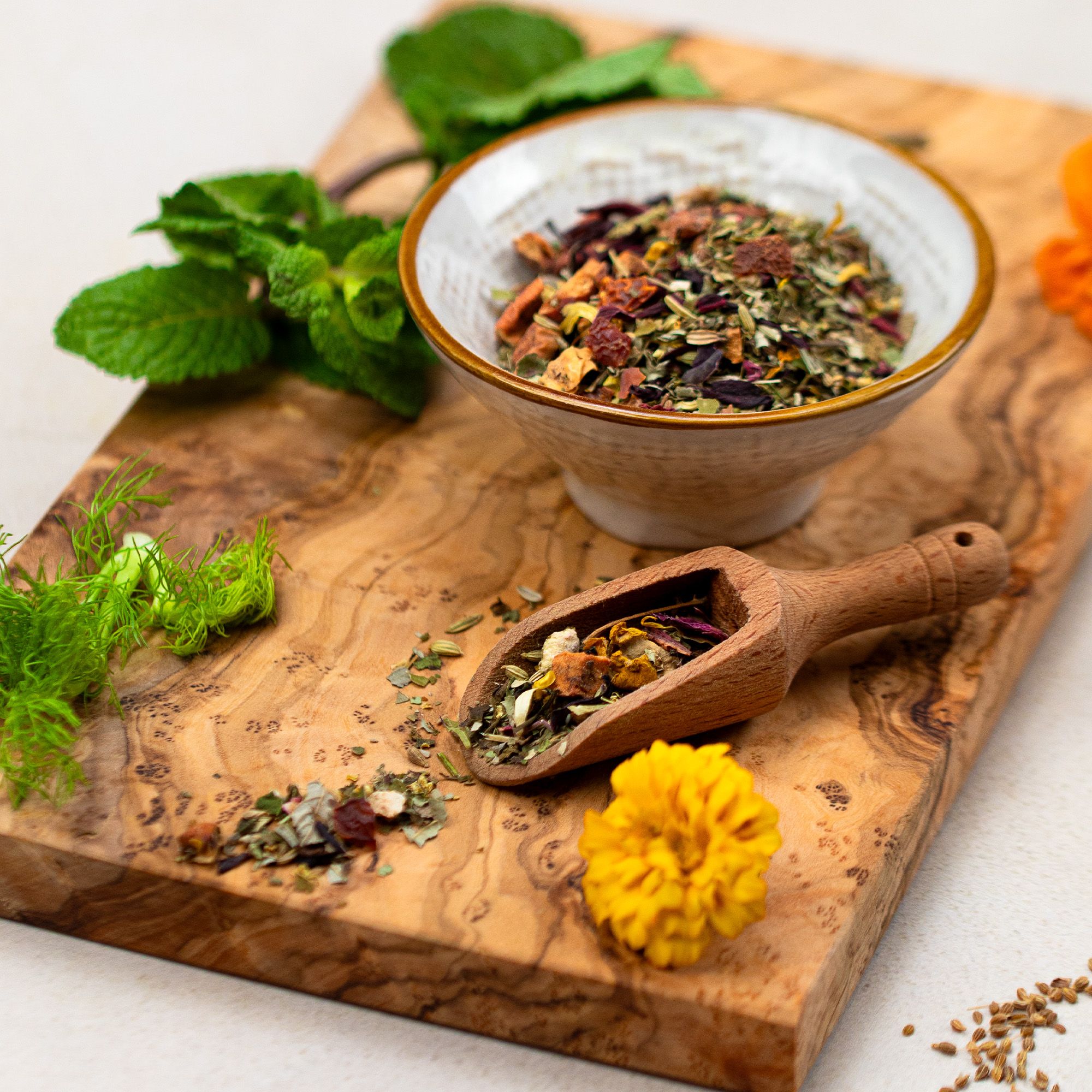 Schale mit Kräutermischung, Holzlöffel. Auf Holzbrett. Minze, Fenchel, Blüten. Helle Schale, brauner Löffel.