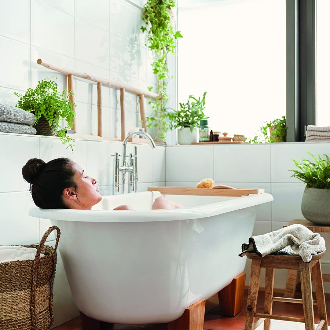 Frau in weißer Badewanne mit Holzleiter und Pflanzen. Badewanne auf Holzbeinen. Graue Handtücher und Holzschemel.