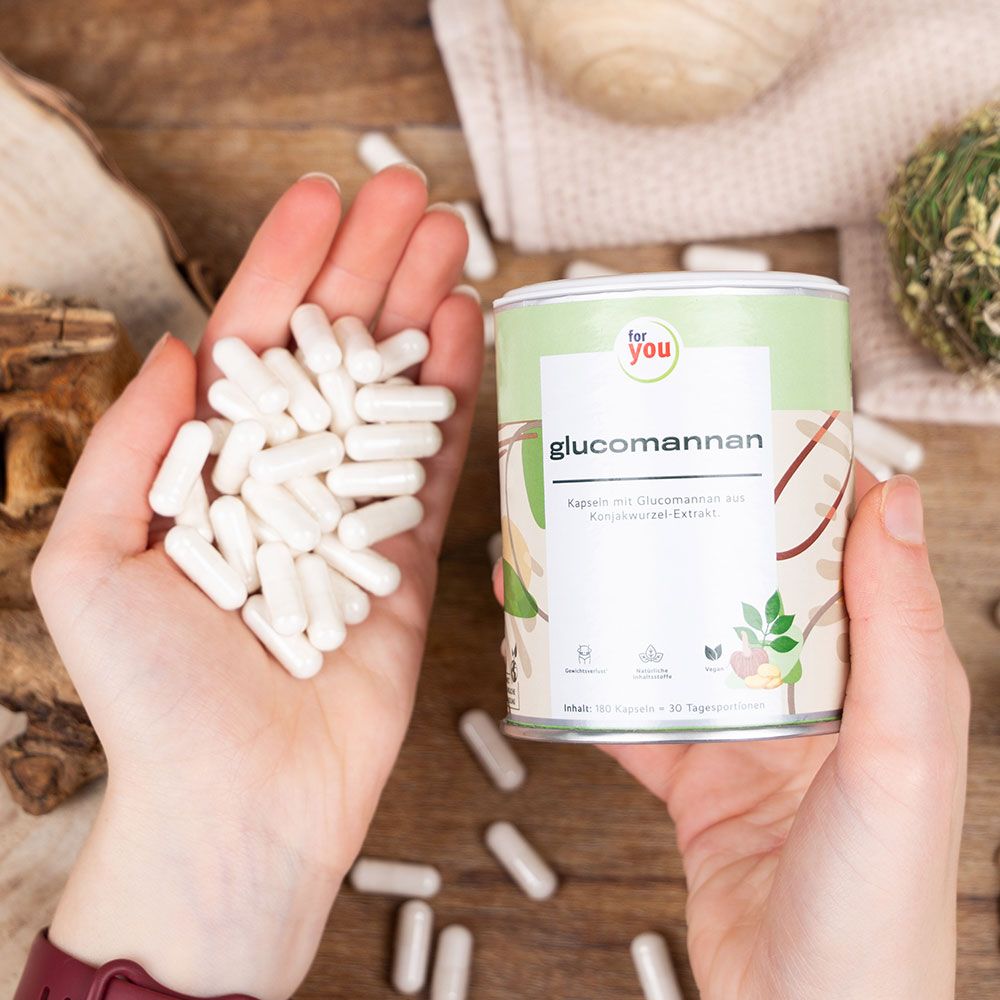 Hände halten weiße Kapseln und Dose "for you Glucomannan". Dose mit grün-weißem Design. Kapseln liegen verstreut auf Holzoberfläche.