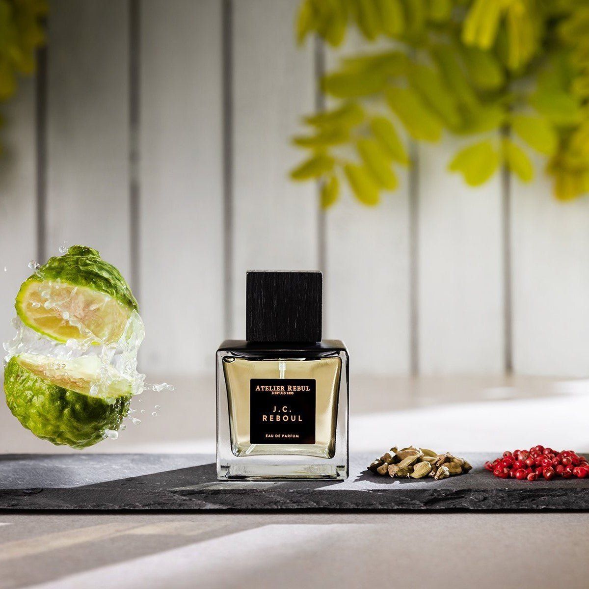 Quadratischer Glasflakon mit schwarzem Verschluss. Aufschrift: Atelier Rebul J.C. Reboul Eau de Parfum. Neben geschnittenen Zitronen.