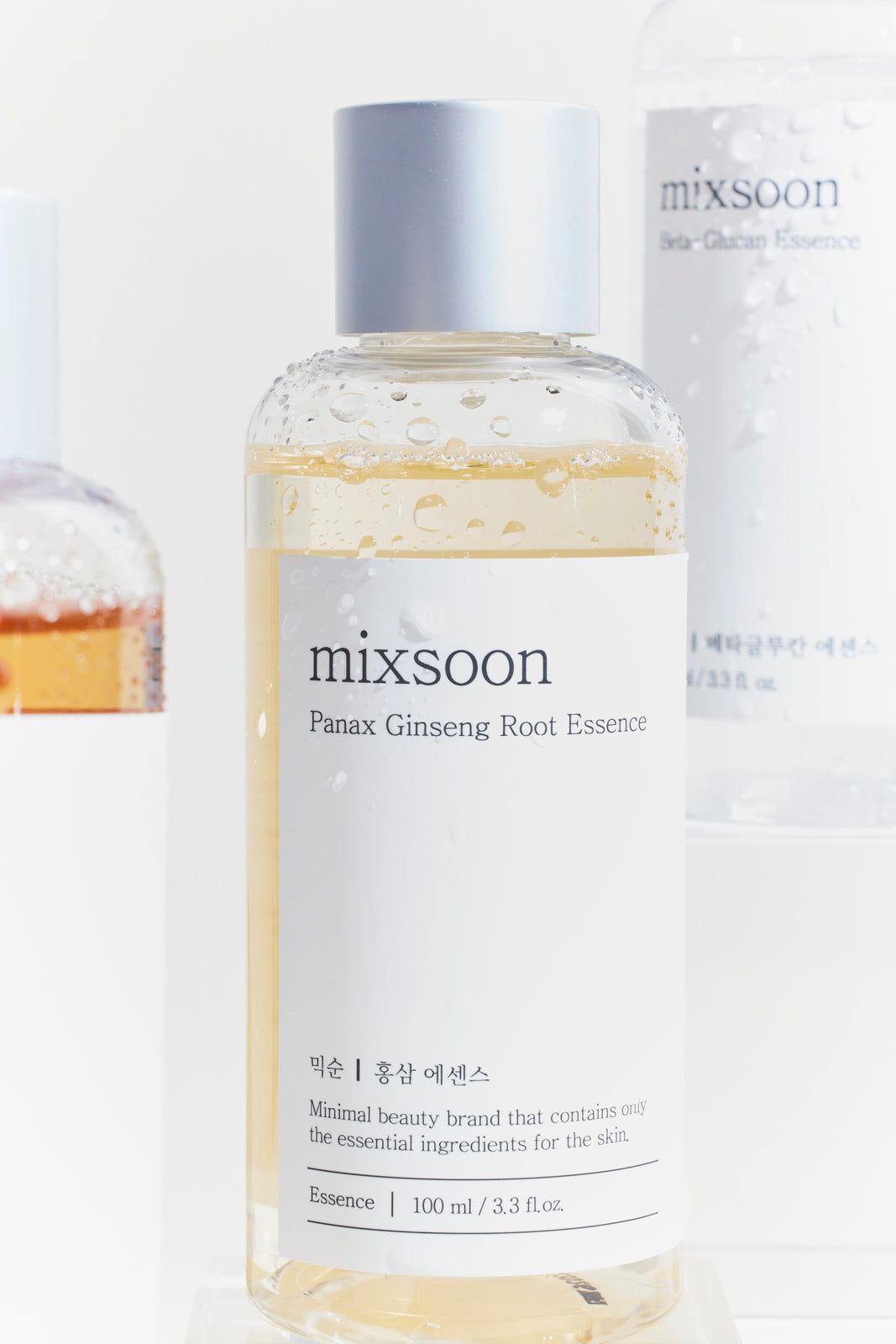 Klare Flasche mit silbernem Deckel. Aufschrift 'mixsoon Panax Ginseng Root Essence'. Mehrere Flaschen im Hintergrund.