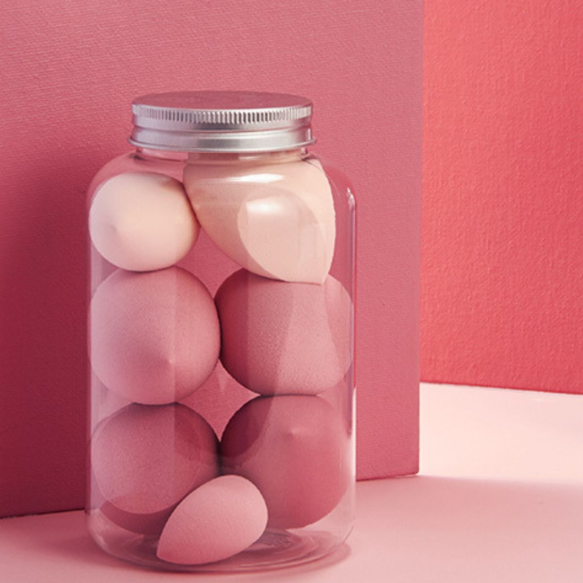 Mehrfarbige Make-up-Schwämme in einer transparenten Dose mit silbernem Deckel. Verschiedene rosa Formen.