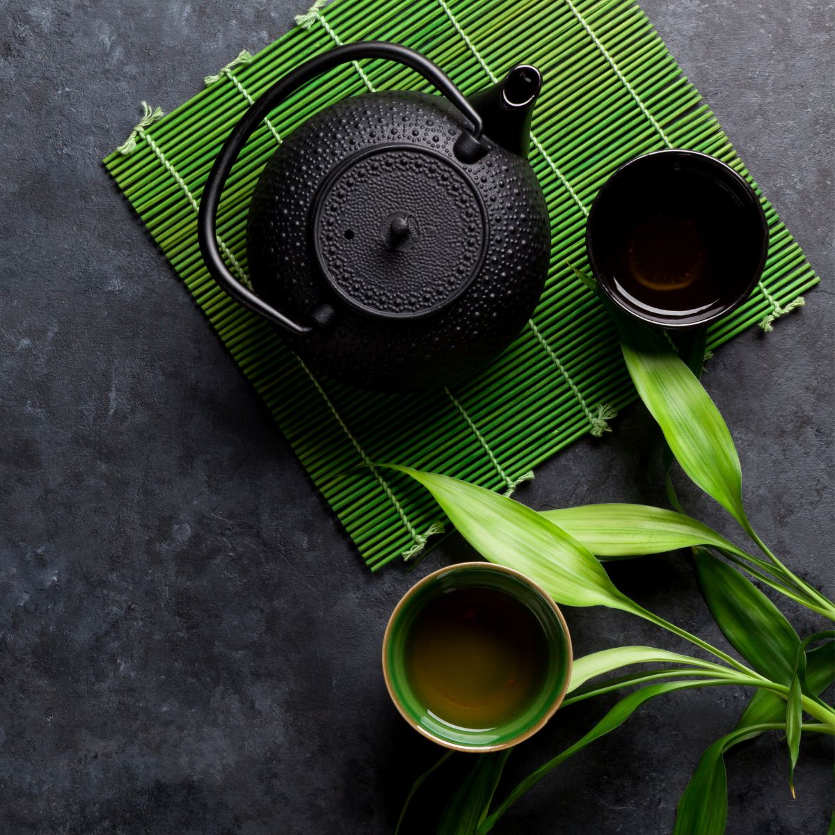 Schwarze Teekanne und zwei Tassen auf Bambusmatte. Grüne Blätter liegen daneben. Draufsicht.