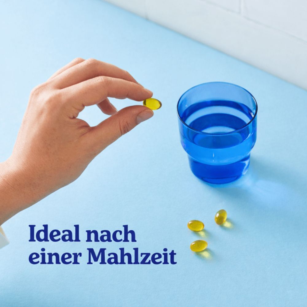Hand hält eine Omega 3 Kapsel über einem blauen Glas mit Wasser.