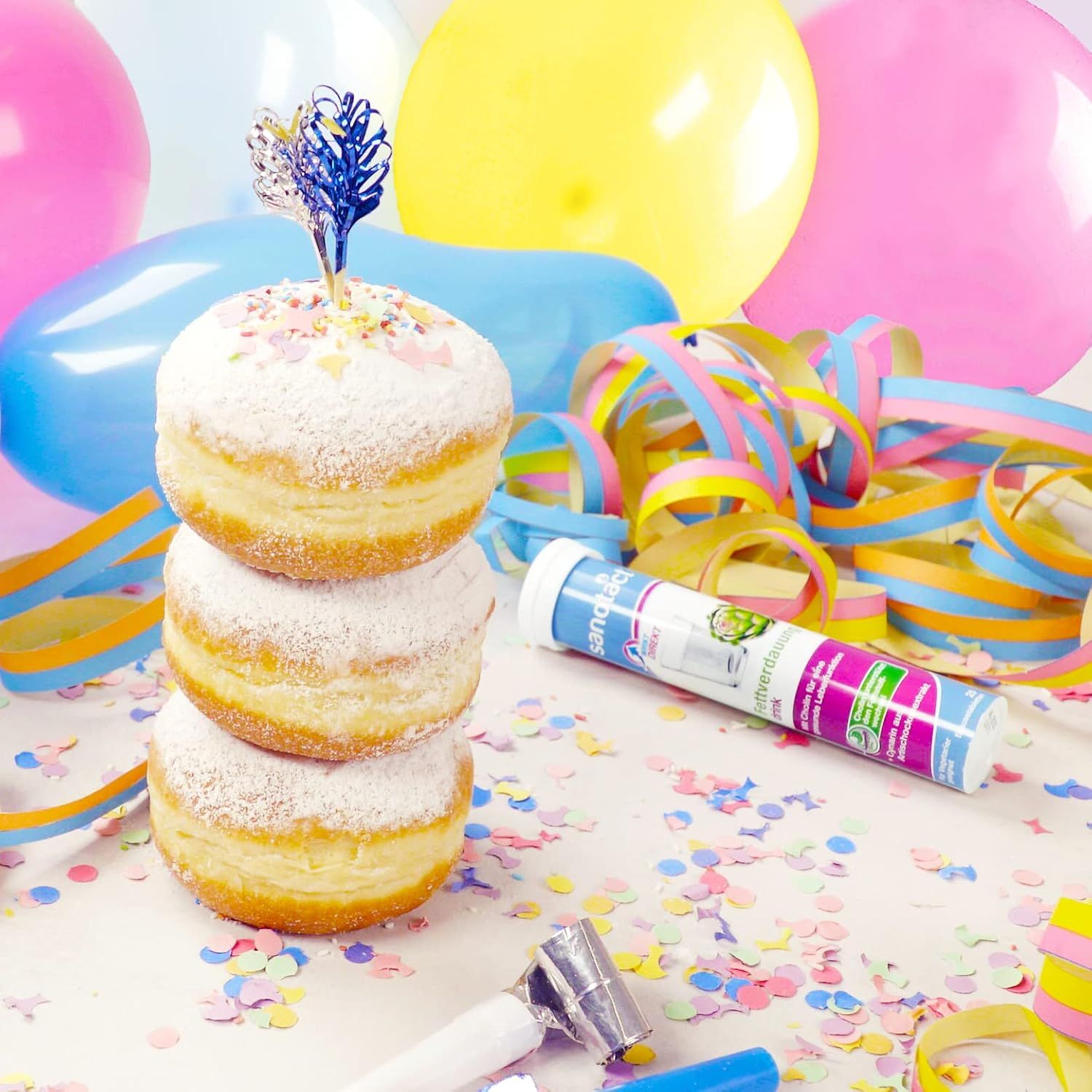 Drei Berliner Pfannkuchen mit Puderzucker. Daneben Konfetti, Luftballons und eine Packung Sanotact Fettverdauung Drink Brausetabletten.