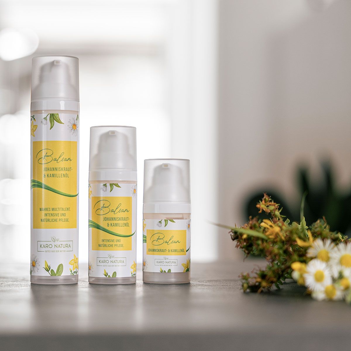 Drei Flaschen mit gelben Etiketten. Aufschrift: Balsam Johanniskraut- & Kamillenöl. Marke: Karo Natura. Daneben Blumen.