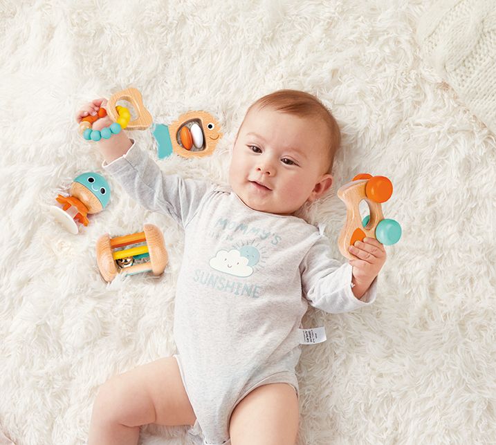 Baby mit Hape Sensorik-Set. Das Kind hält fünf Holzspielzeuge mit bunten Elementen. Das Kind liegt auf einer weißen Decke.