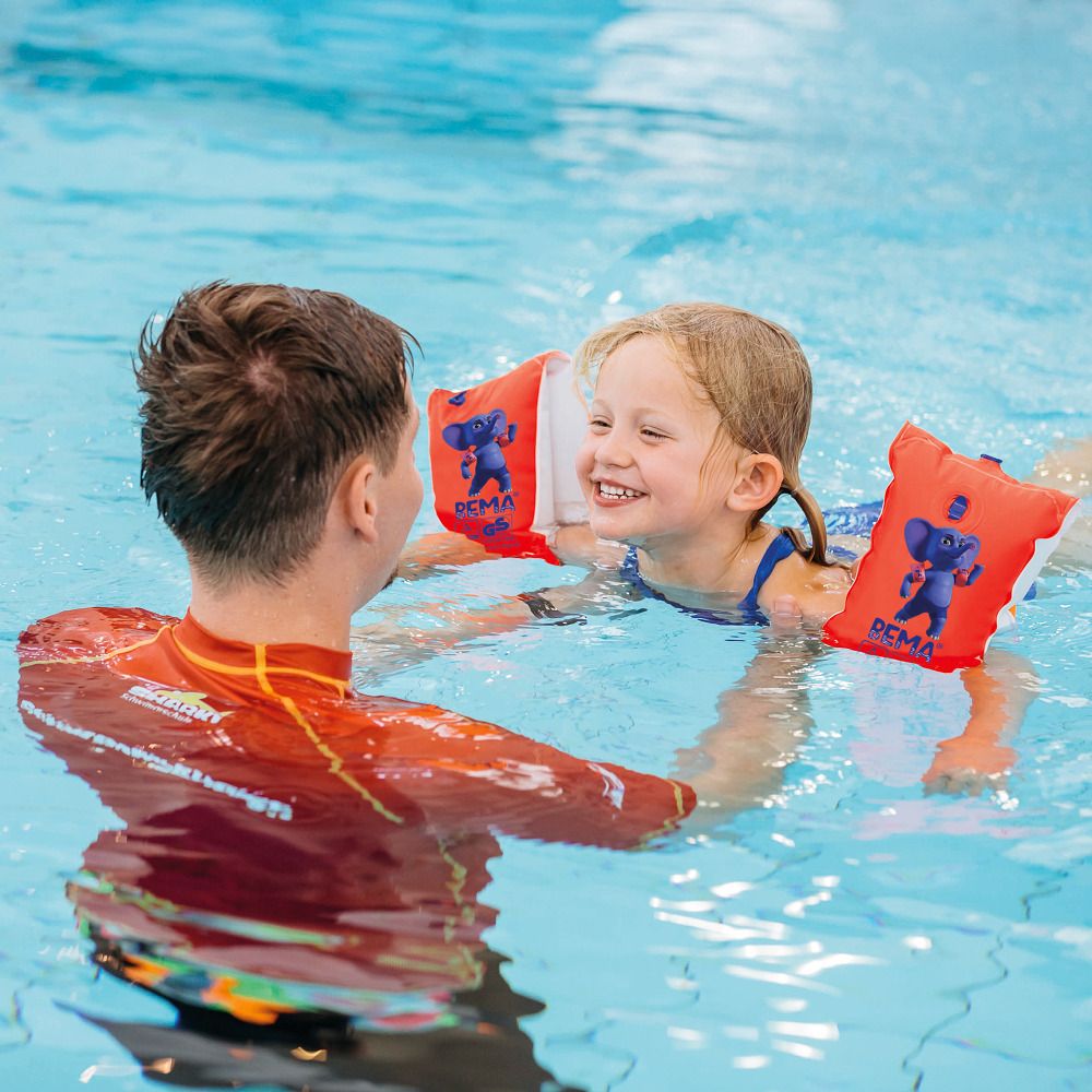 Kind mit BEMA-Schwimmflügeln im Wasser. Ein Mann hilft dem Kind. Die Schwimmflügel sind orange mit einem blauen Elefanten.