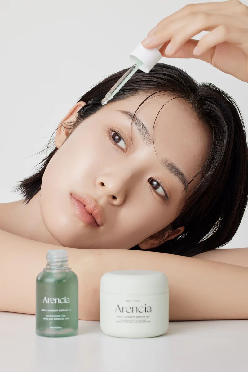 Frau mit Serumflasche und Creme. Aufschrift: Aarencia, Holy Hyssop Serum 12, Niacinamide 10% + Vita Ace Complex 12%. Creme: Aarencia.