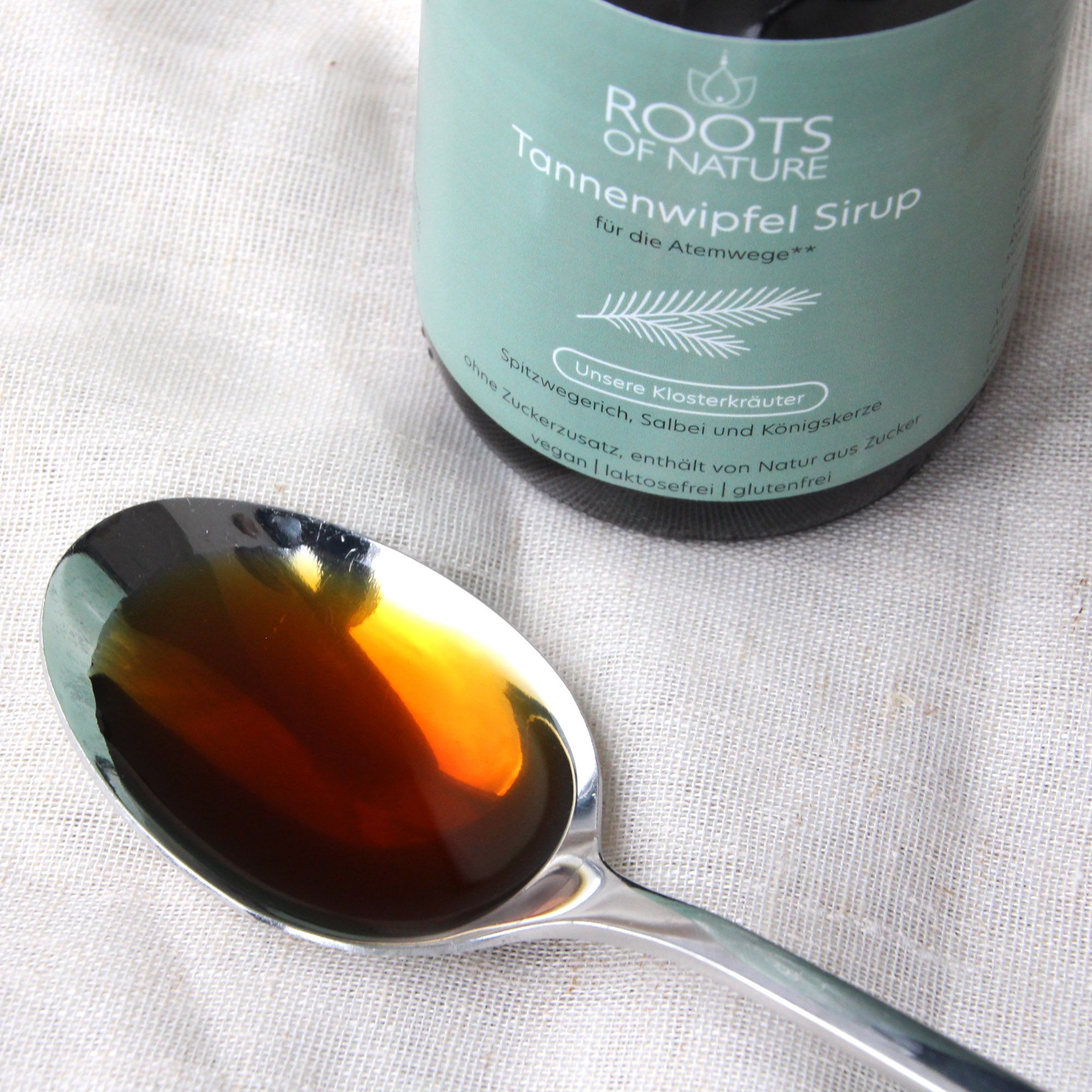 Löffel mit brauner Flüssigkeit, daneben eine Flasche ROOTS OF NATURE Tannensirup. Aufschrift: Tannensirup für die Atemwege.