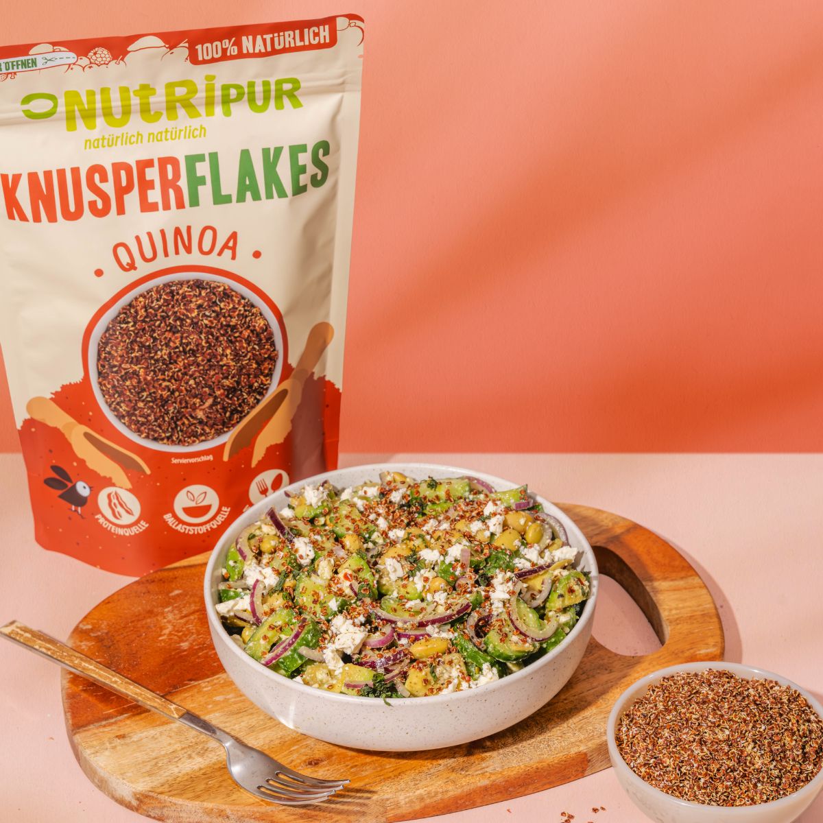 Beutel NutriPur Knusperflakes Quinoa neben Salat mit Flakes. Schüssel mit Flakes und eine Schüssel mit Salat auf Holzbrett.