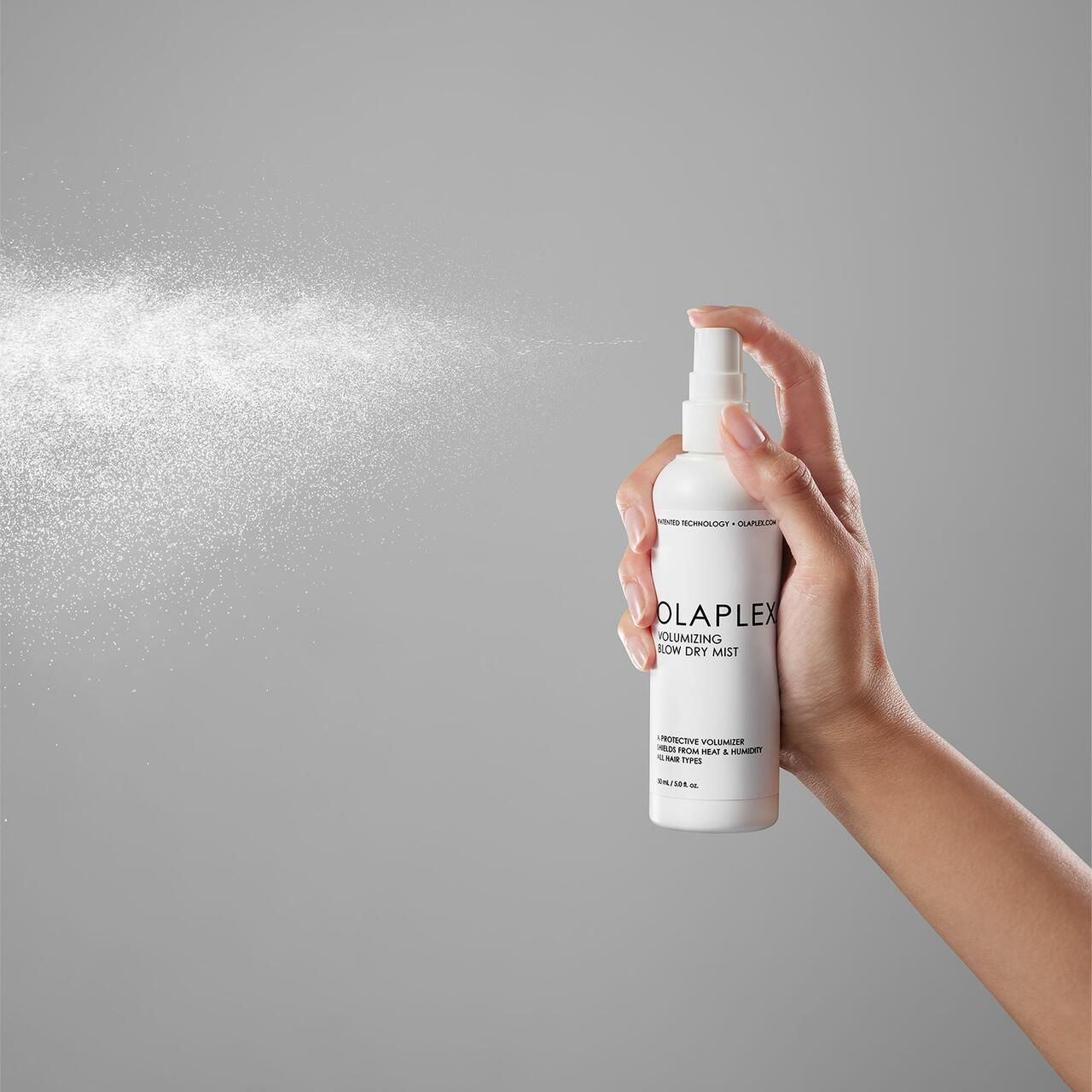 Hand hält weiße Sprühflasche. Sprühnebel sichtbar. Olaplex-Logo und Produktbezeichnung auf der Flasche.