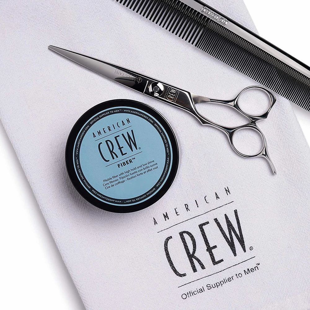 Dunkelblaue Dose mit "AMERICAN CREW FIBER"-Aufschrift, Schere, Kamm und Handtuch. Aufschrift "Official Supplier to Men".