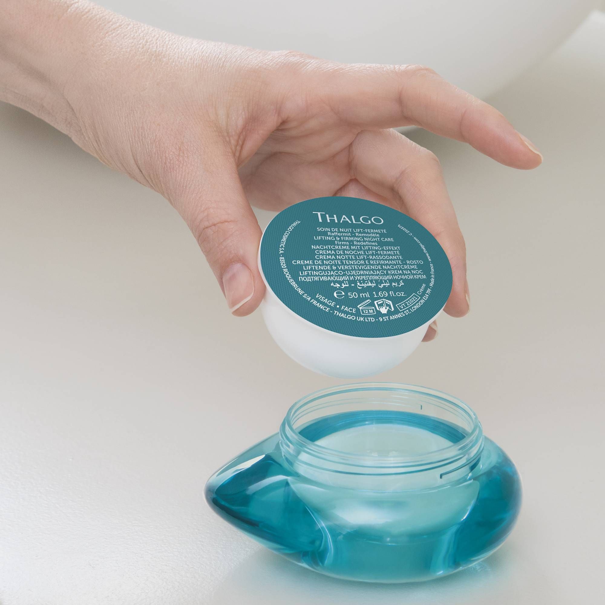 Hand öffnet Cremebehälter. Weißer Behälter mit blauem Deckel. Aufschrift: THALGO. Offener, blauer Glasbehälter.