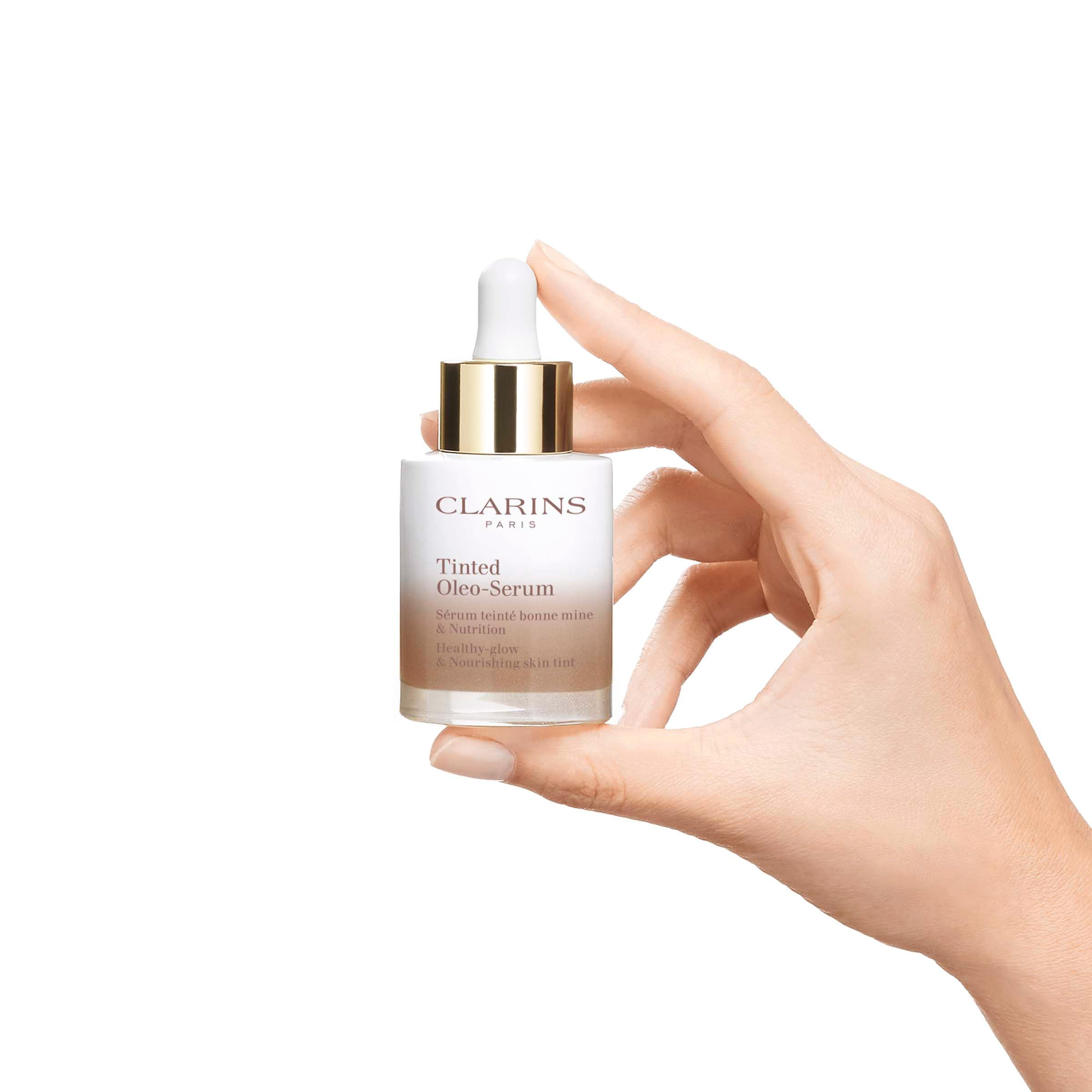 Hand hält Clarins Tinted Oleo-Serum Flasche. Weißes Glas, goldener Deckel, Produktbezeichnung.