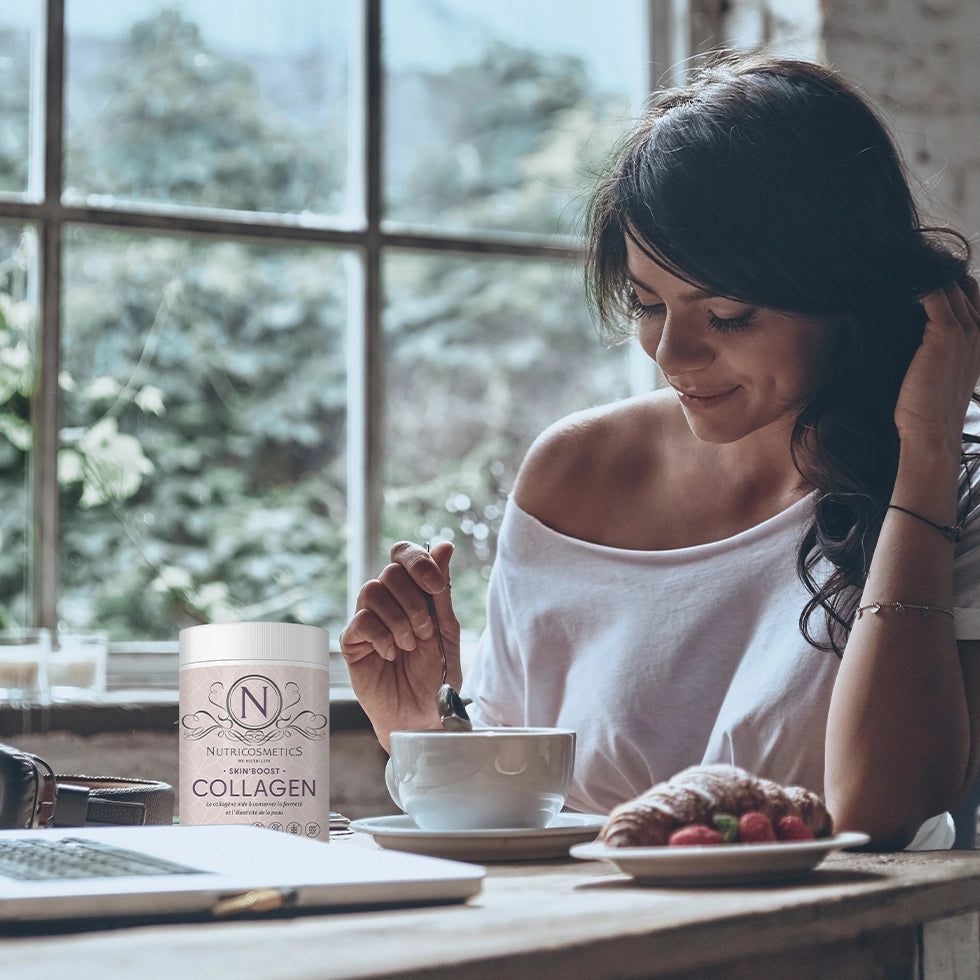 Frau sitzt am Tisch, Dose mit Collagen neben Tasse und Croissant. Fenster im Hintergrund.