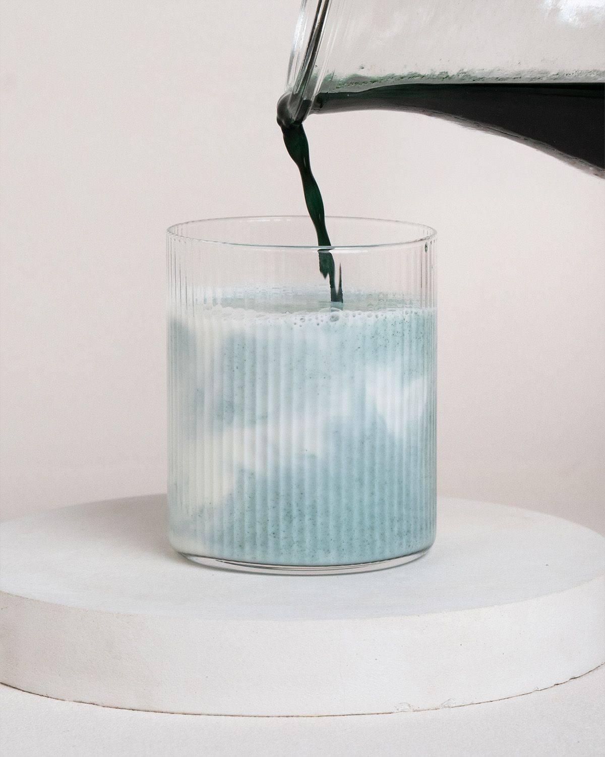 Grünes Getränk wird in ein geriffeltes Glas gegossen, das auf einem weißen Podest steht. Der Inhalt des Glases ist blau.