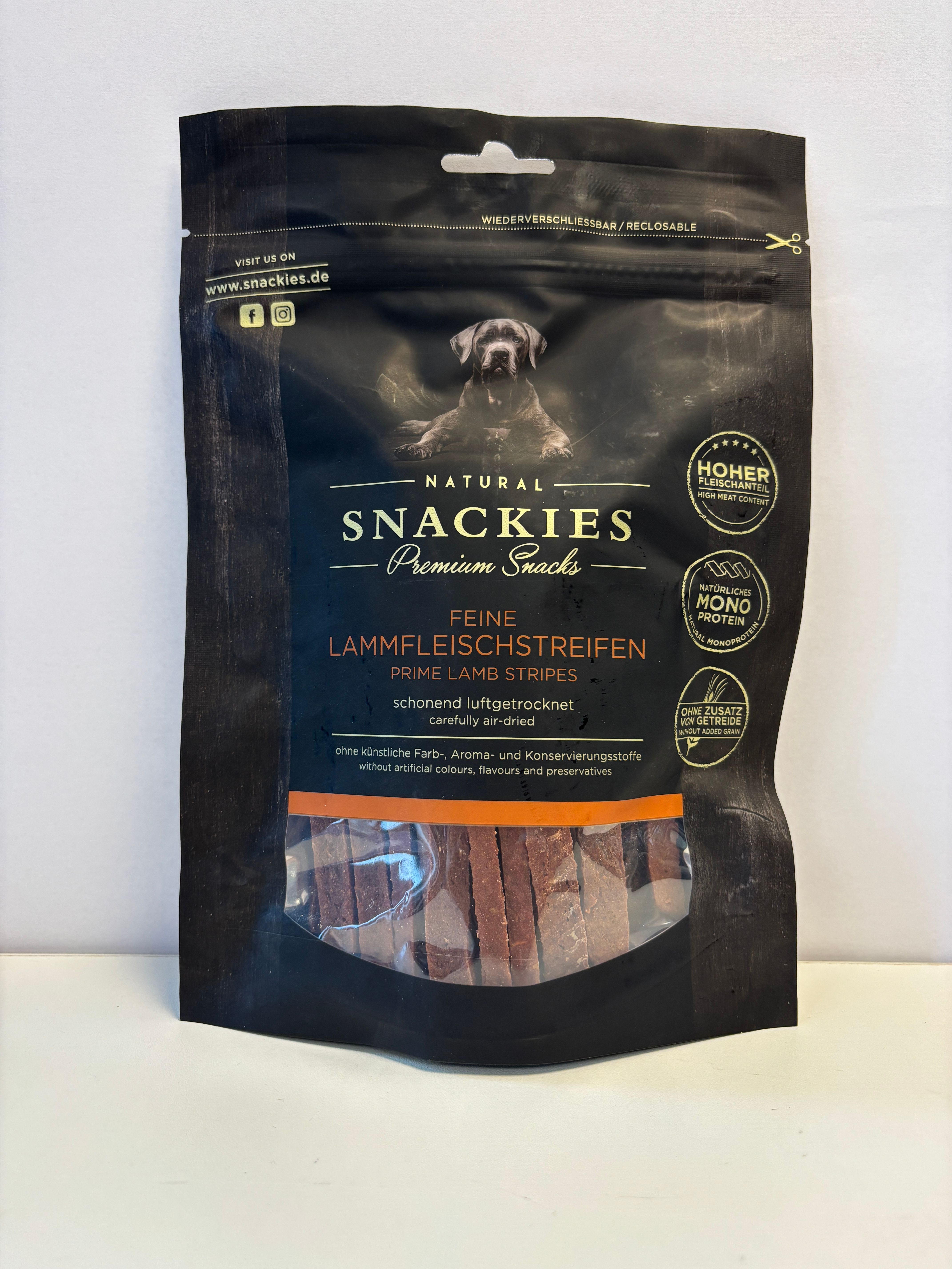 Verpackung mit Snackies Feine Lammfleischstreifen. Schwarze Tüte mit Produktnamen, Hund und Produktstreifen.