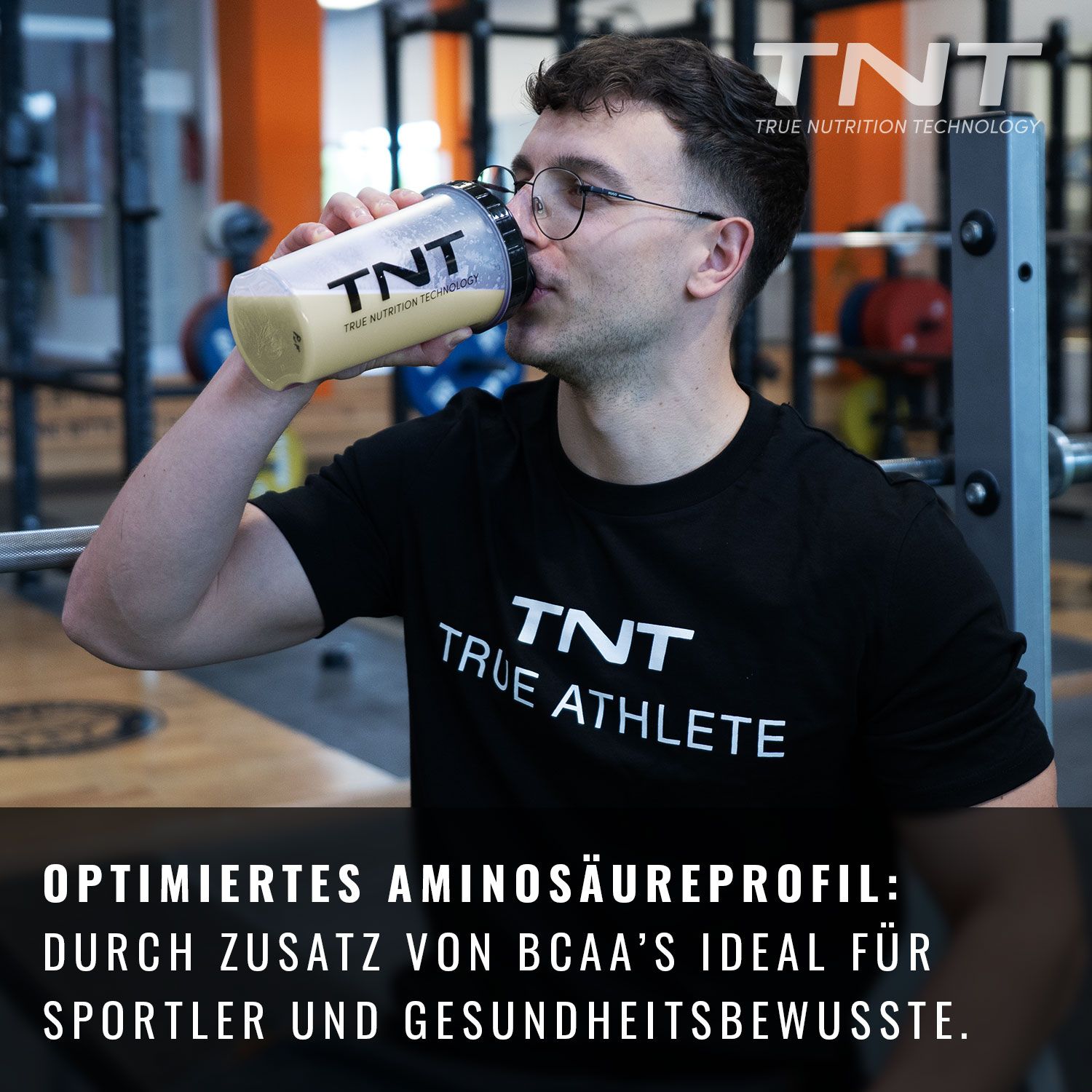 Mann trinkt Protein-Shake aus einem Shaker. Auf dem Shaker steht TNT. Im Hintergrund Fitnessstudio.