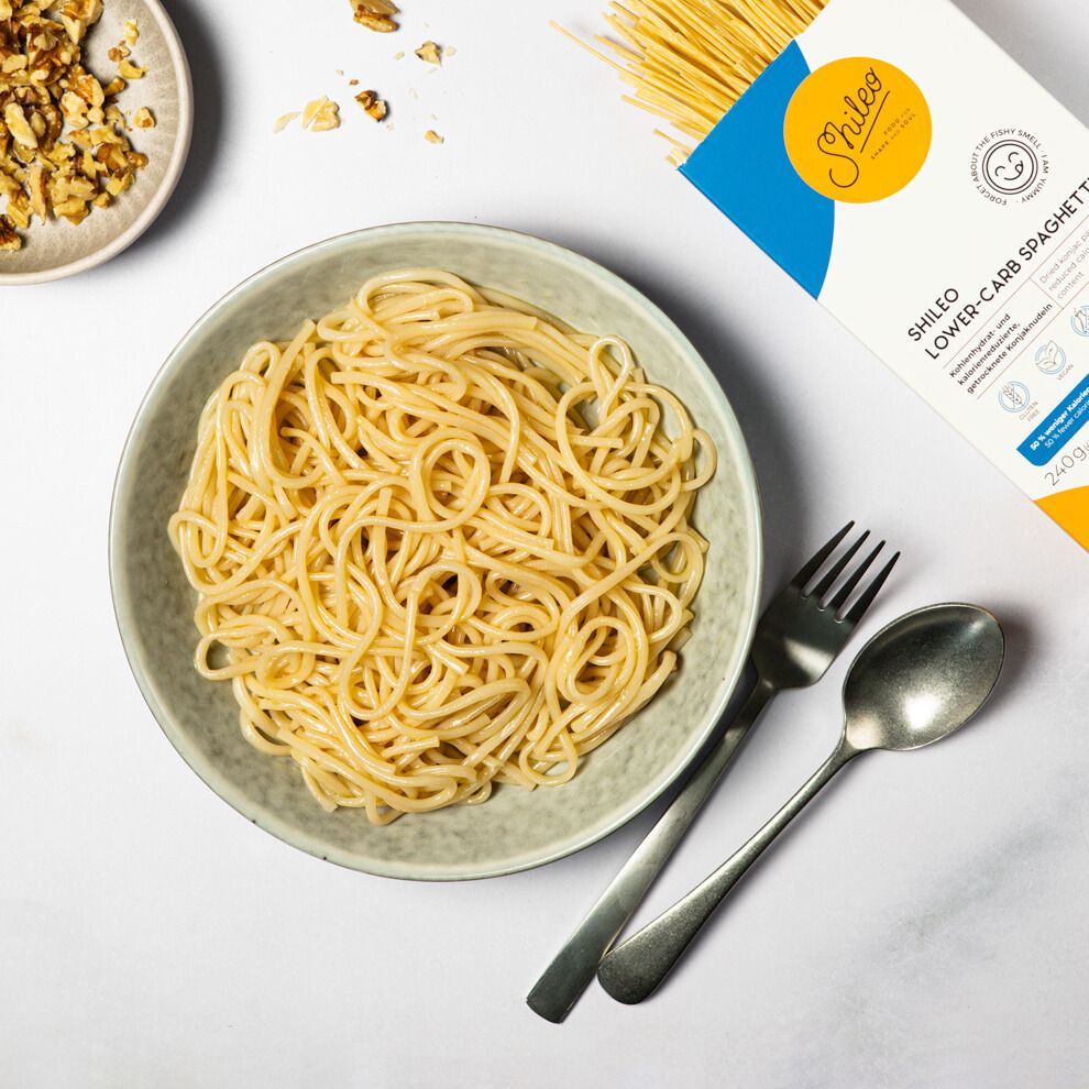 Teller mit Spaghetti. Gabel und Löffel daneben. Aufdrucke: Logo, Produktname. Auf dem Teller: Spaghetti. Auf dem Tisch: Walnüsse.